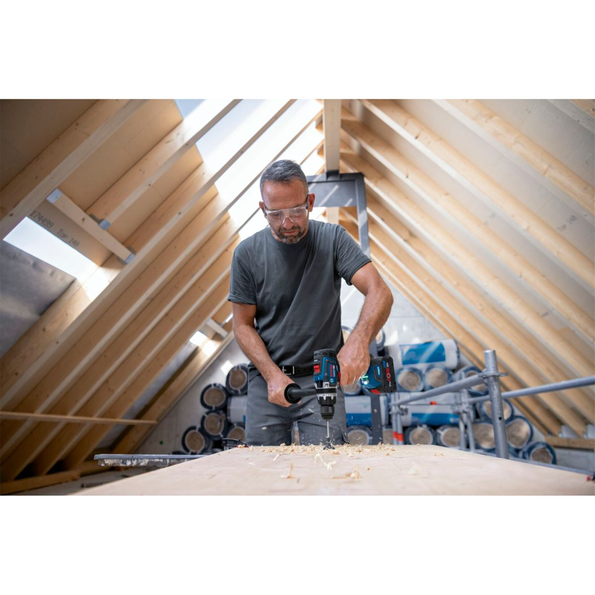 Mężczyzna wierci w drewnie w drewnianej konstrukcji. Ma okulary ochronne i szarą koszulę. Widoczne trociny.