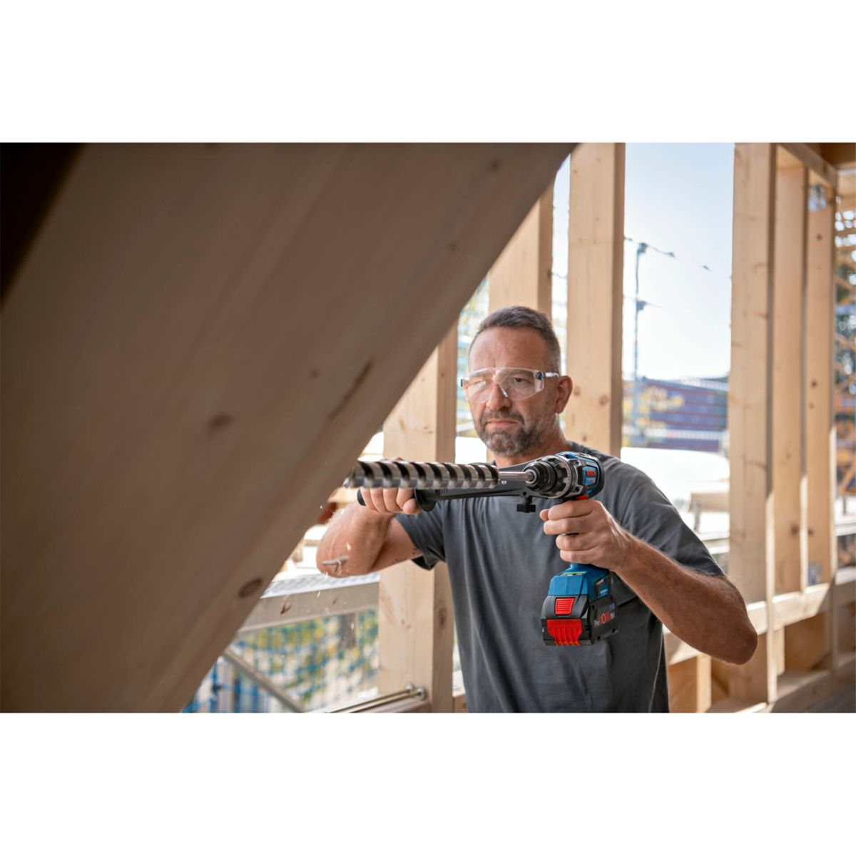 Mężczyzna wierci drewno wiertarką. Ma na sobie okulary ochronne. Drewniana rama i plac budowy w tle.