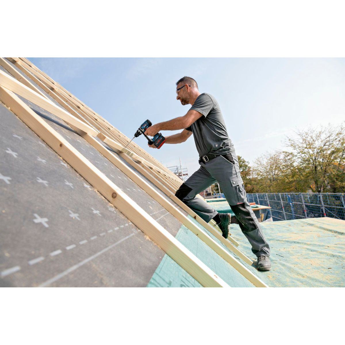 Robotnik wierci w ramie dachu. Ma na sobie okulary i szare ubranie robocze. Niebo jest niebieskie, a w tle widać drzewa.