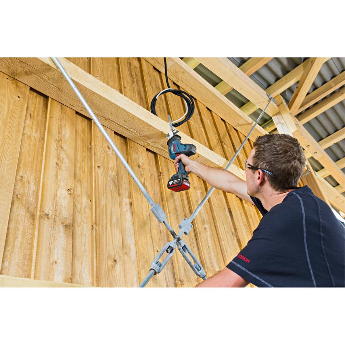 Mężczyzna używa piły elektrycznej do cięcia drewnianych belek. Ma okulary i czarną koszulę. Piła obok kabli elektrycznych i konstrukcji.