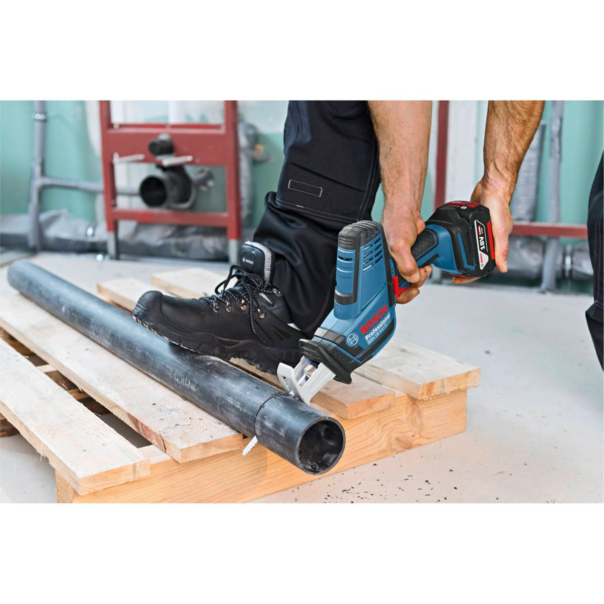 Robotnik używa niebieskiej piły Bosch do cięcia czarnej rury. Rura spoczywa na drewnianej palecie. Robotnik nosi czarne buty robocze.