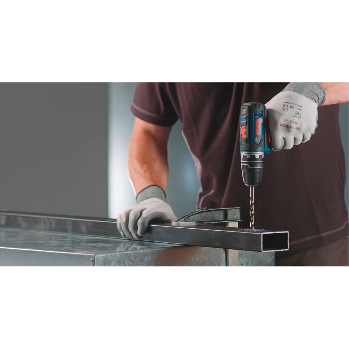 Osoba wierci otwór w metalowej belce wiertarką Bosch. Mają na sobie rękawice i ciemną koszulę. Tło to szara ściana.