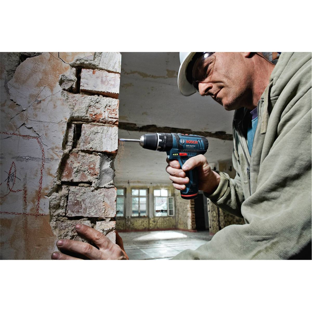 Mężczyzna w kasku wierci w ceglanym murze. Używa niebiesko-czarnej wiertarki. Tło przedstawia pokój z oknami.