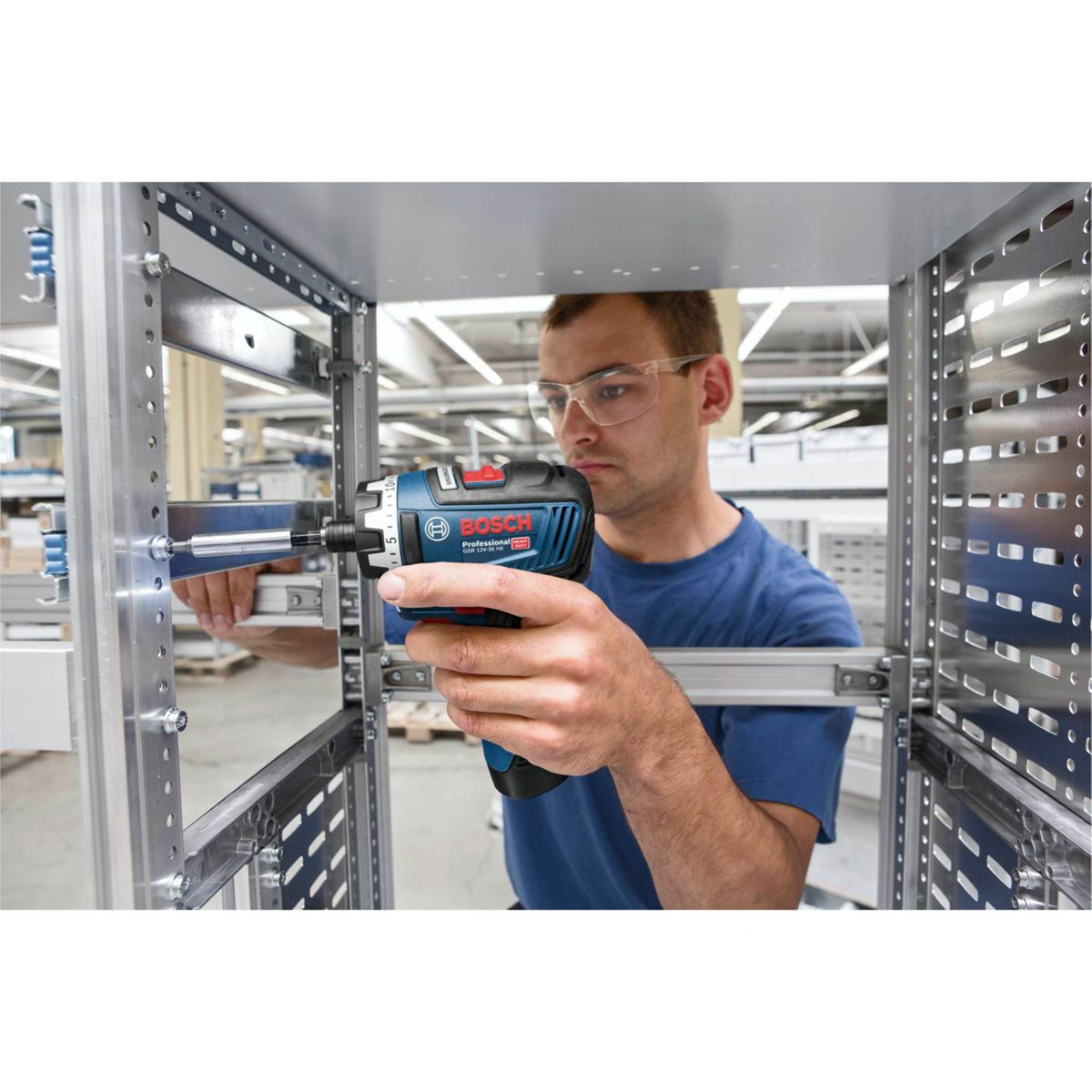 Mężczyzna w okularach ochronnych używa wiertarki Bosch na metalowej ramie. Wiertarka jest niebiesko-czarna. Tło to fabryka.