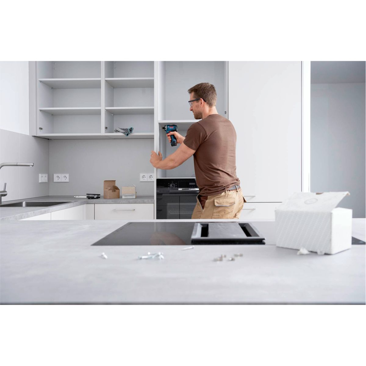 Mężczyzna w brązowej koszuli i okularach wierci drzwi szafki w nowoczesnej kuchni. Białe pudełko stoi na blacie.