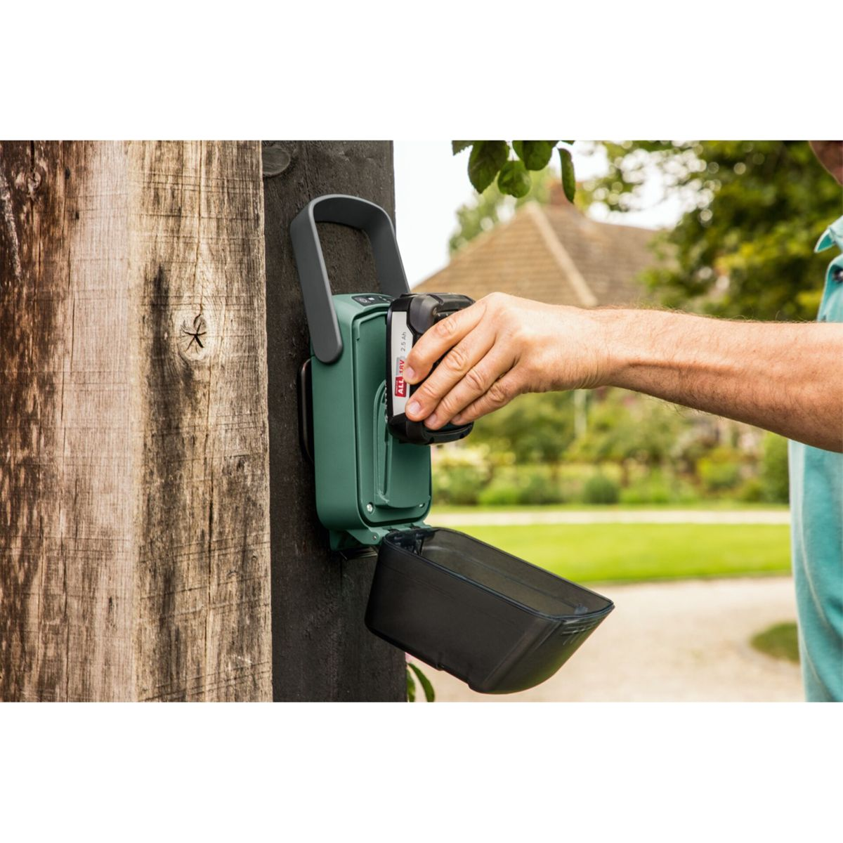 Osoba wkłada baterię do zielonego urządzenia przymocowanego do drewnianego słupa. Czarny pojemnik jest otwarty pod urządzeniem.