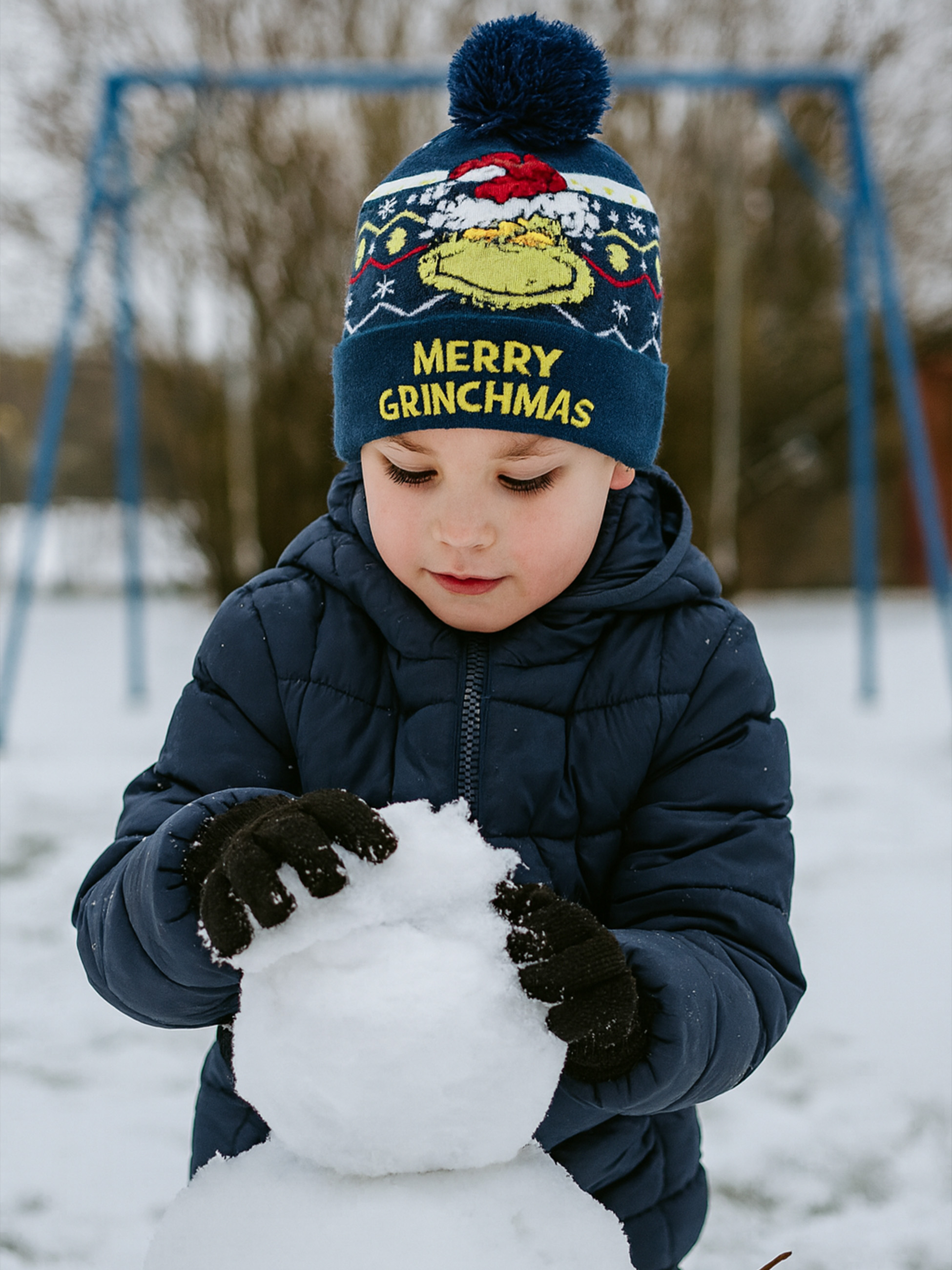 Ein Kind in einer blauen Jacke und Mütze baut einen Schneemann im Schnee. Auf der Mütze steht 'Merry Grinchmas'.