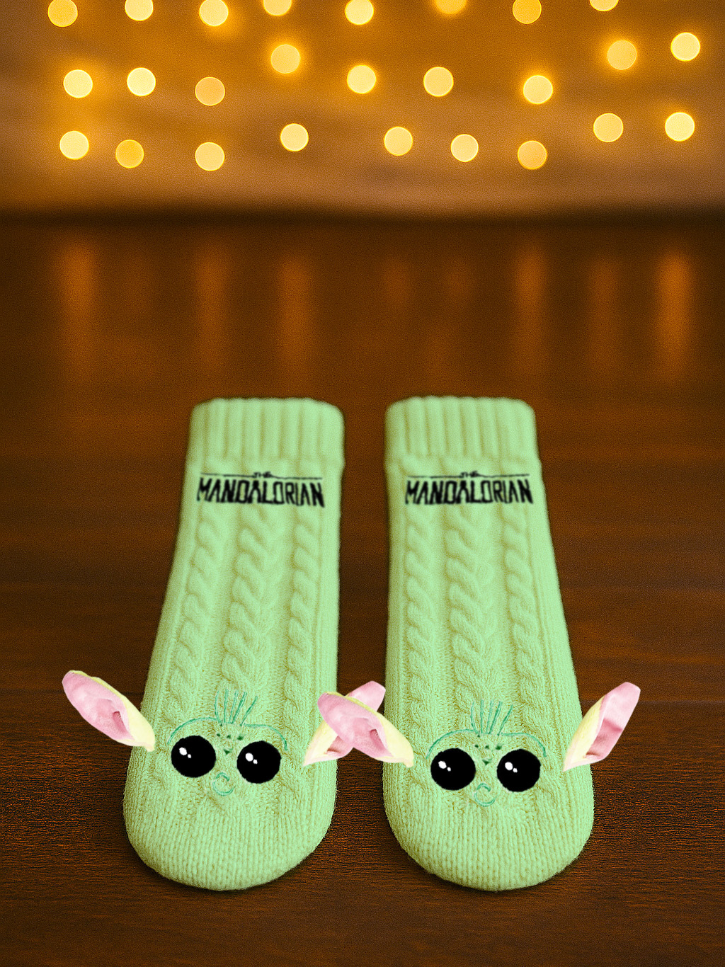 Zwei hellgrüne Socken mit Grogu-Gesichtern, Text 'Mandalorian', auf Holz, mit Bokeh-Lichtern im Hintergrund.