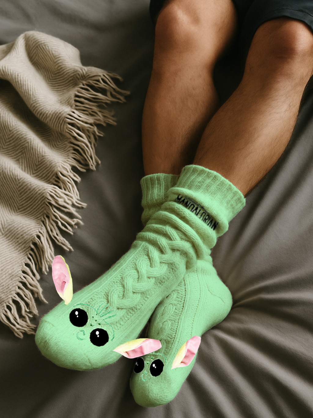Beine einer Person mit grünen Socken, die als Zeichentrickfigur gestaltet sind. Decke links, Beine gekreuzt auf einem grauen Bett.