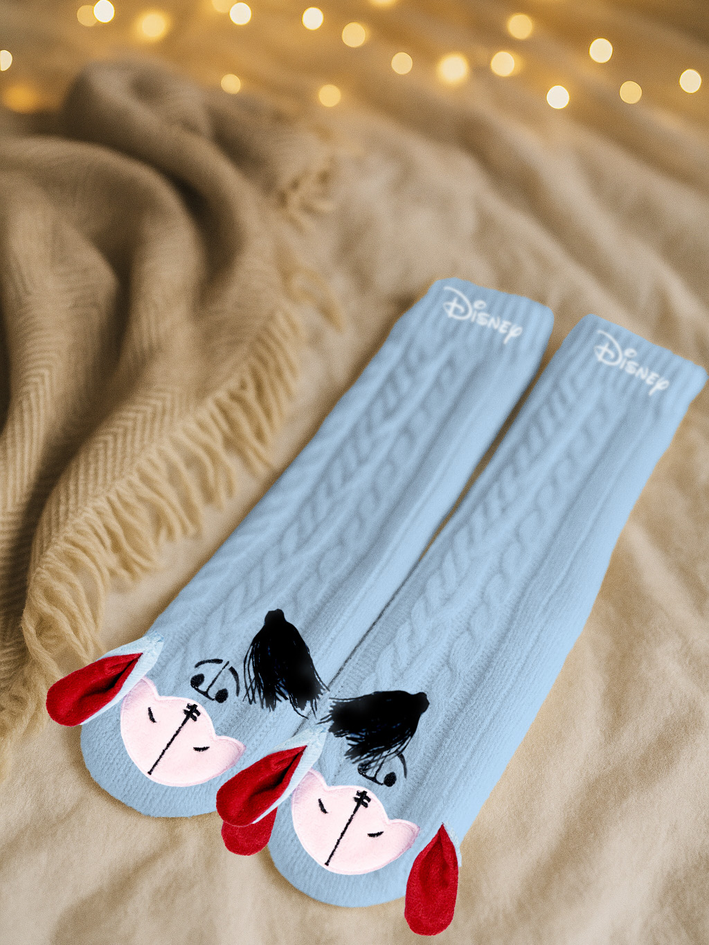 Zwei hellblaue Disney-Socken mit einem Cartoon-Gesicht, roten Ohren, schwarzen Haaren und dem Disney-Logo liegen auf einer beigen Decke.