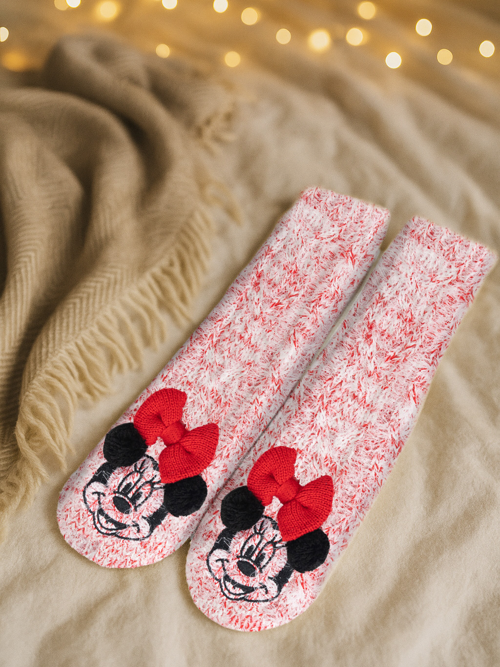 Zwei rot-weiß gestreifte Minnie-Mouse-Socken mit roten Schleifen auf einer beigen Decke. Lichter im Hintergrund.