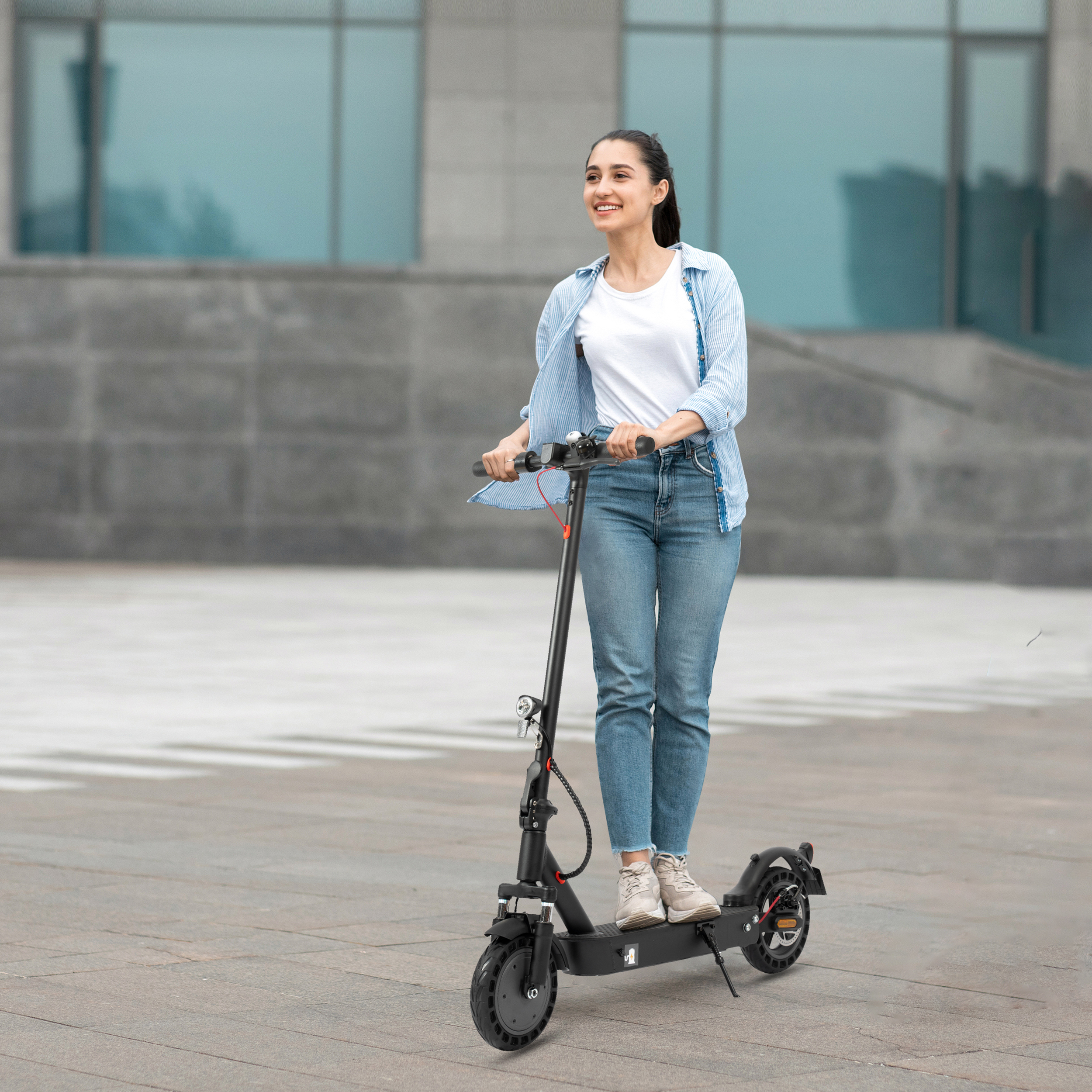 Kobieta jedzie czarną hulajnogą elektryczną. Uśmiecha się, ma na sobie dżinsy i koszulę. Tłem jest budynek.