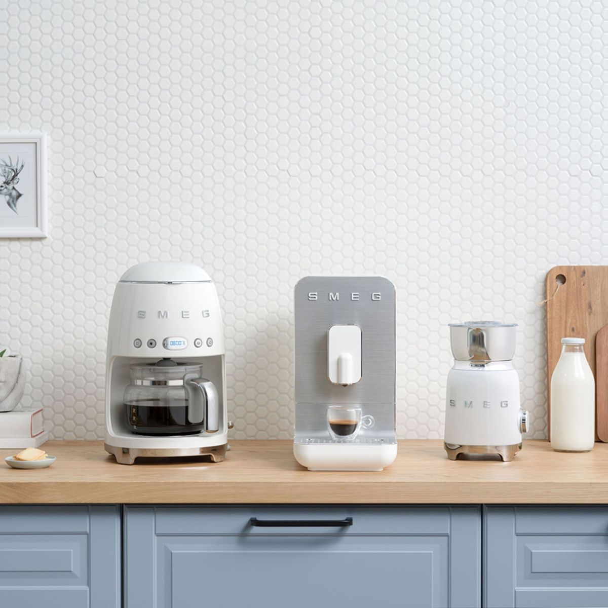 Blat kuchenny z białymi urządzeniami: ekspres do kawy, ekspres do espresso i młynek. Obecna jest również butelka mleka i drewniana deska do krojenia.