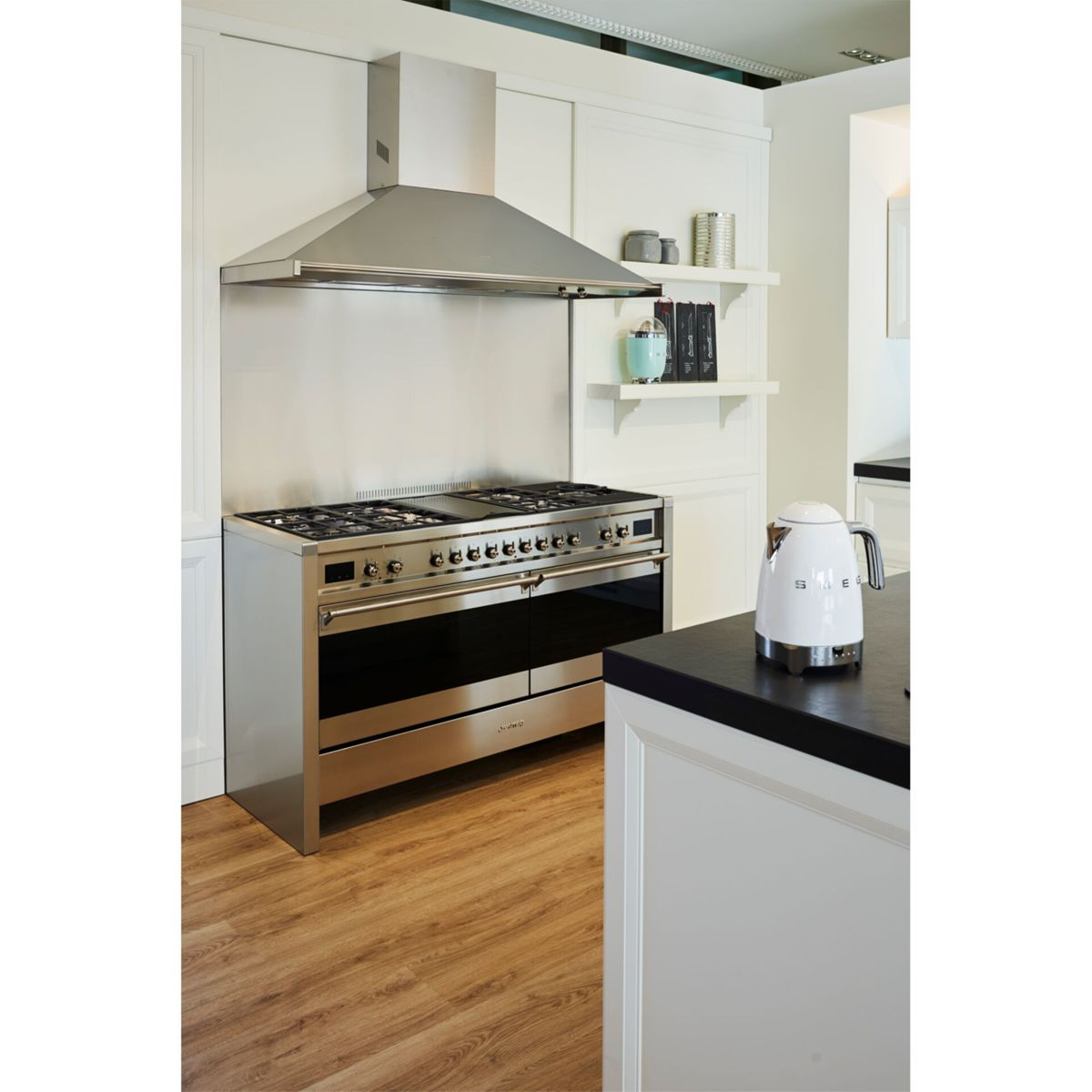 Nowoczesna kuchnia ze stalową kuchenką, okapem i białym czajnikiem na czarnym blacie. Drewniana podłoga i białe szafki.