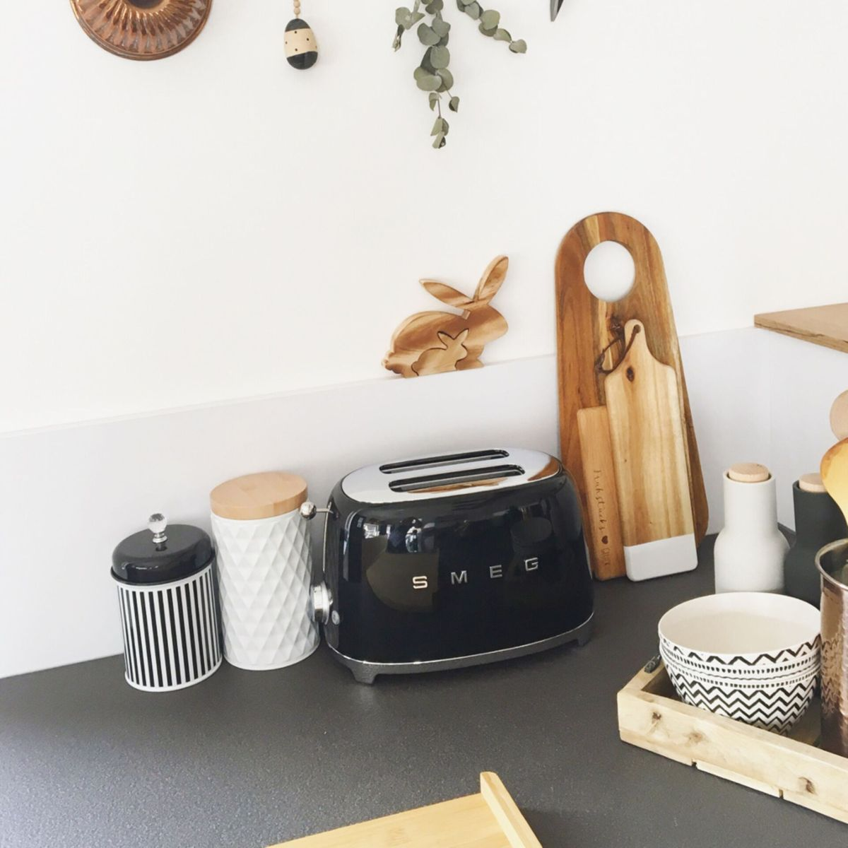 Blat kuchenny z czarnym tosterem, drewnianymi deskami do krojenia, pojemnikami i dekoracyjnym drewnianym zającem. Tło jest białe.
