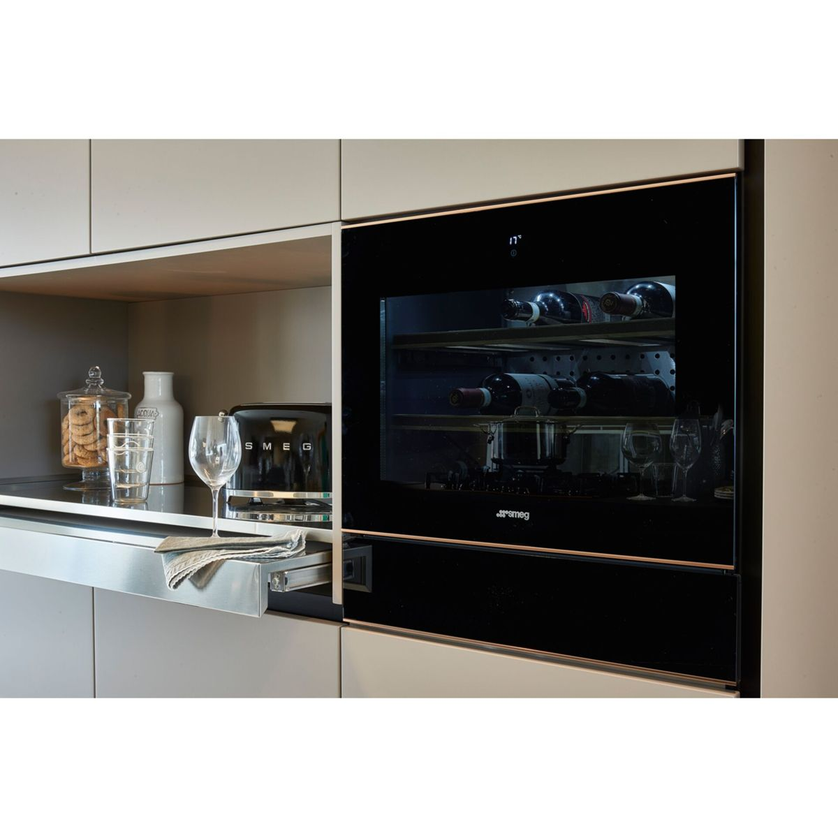 Scena kuchenna z chłodziarką do wina, tosterem, szklankami i słoikami na półce. Butelki wina są widoczne wewnątrz chłodziarki.