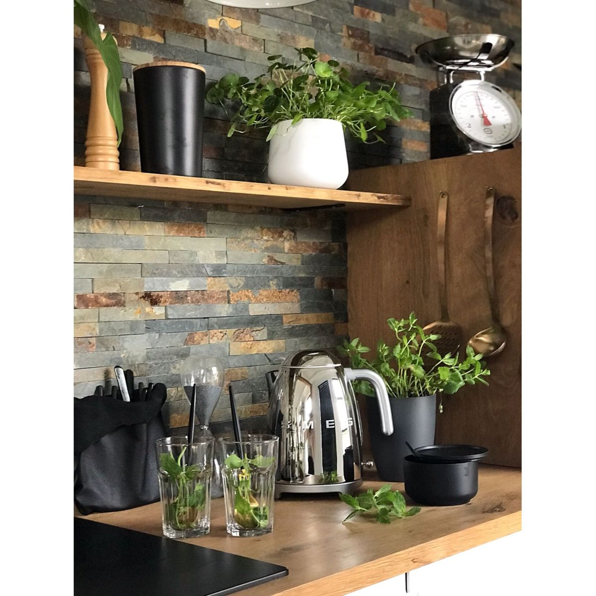 Scena kuchenna z ziołami, szklankami, czajnikiem i wagą. Kamienna ściana w tle.