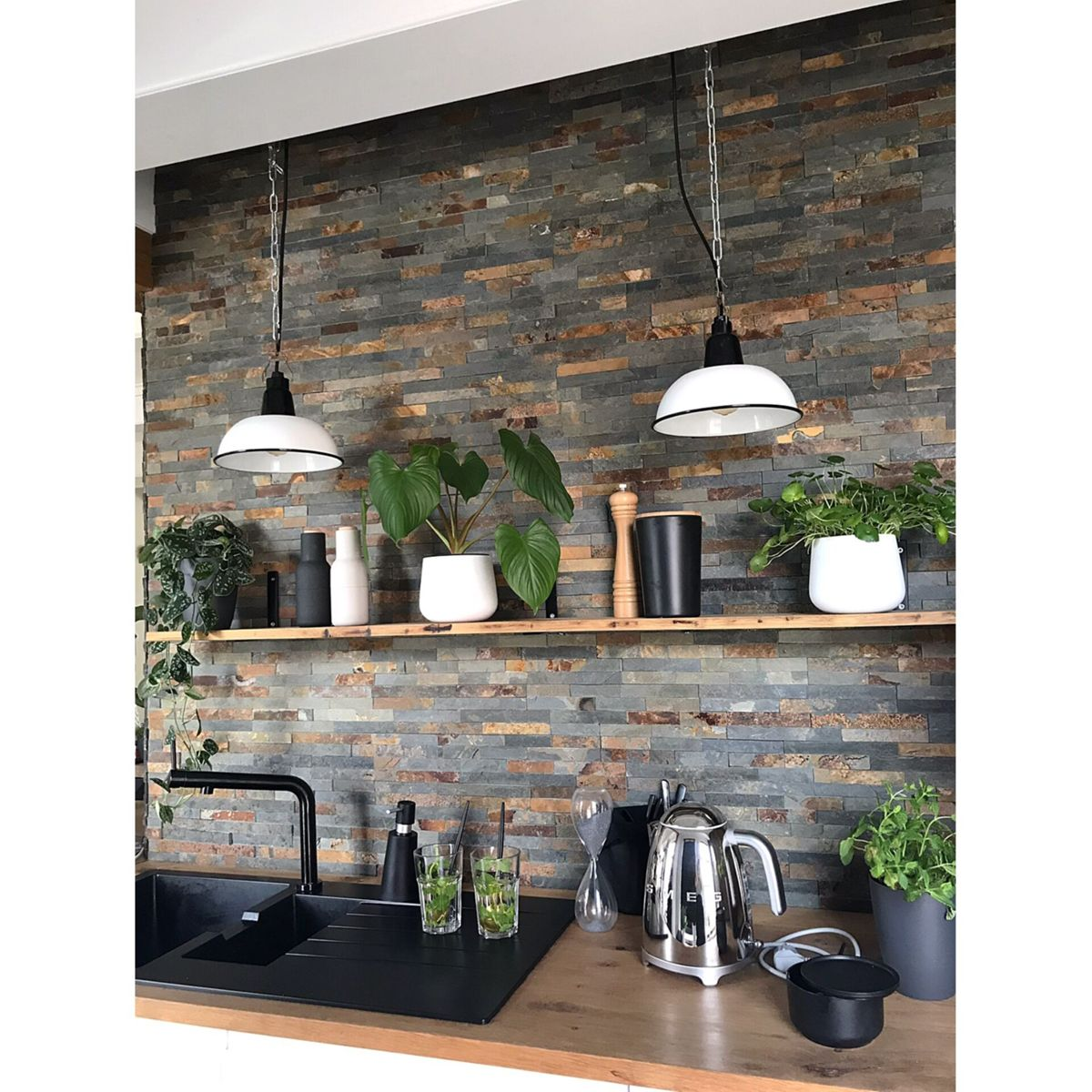 Scena kuchenna z czarnym zlewem, kamienną ścianą, półkami z roślinami i dekoracjami oraz wiszącymi białymi lampami. Czajnik stoi na blacie.