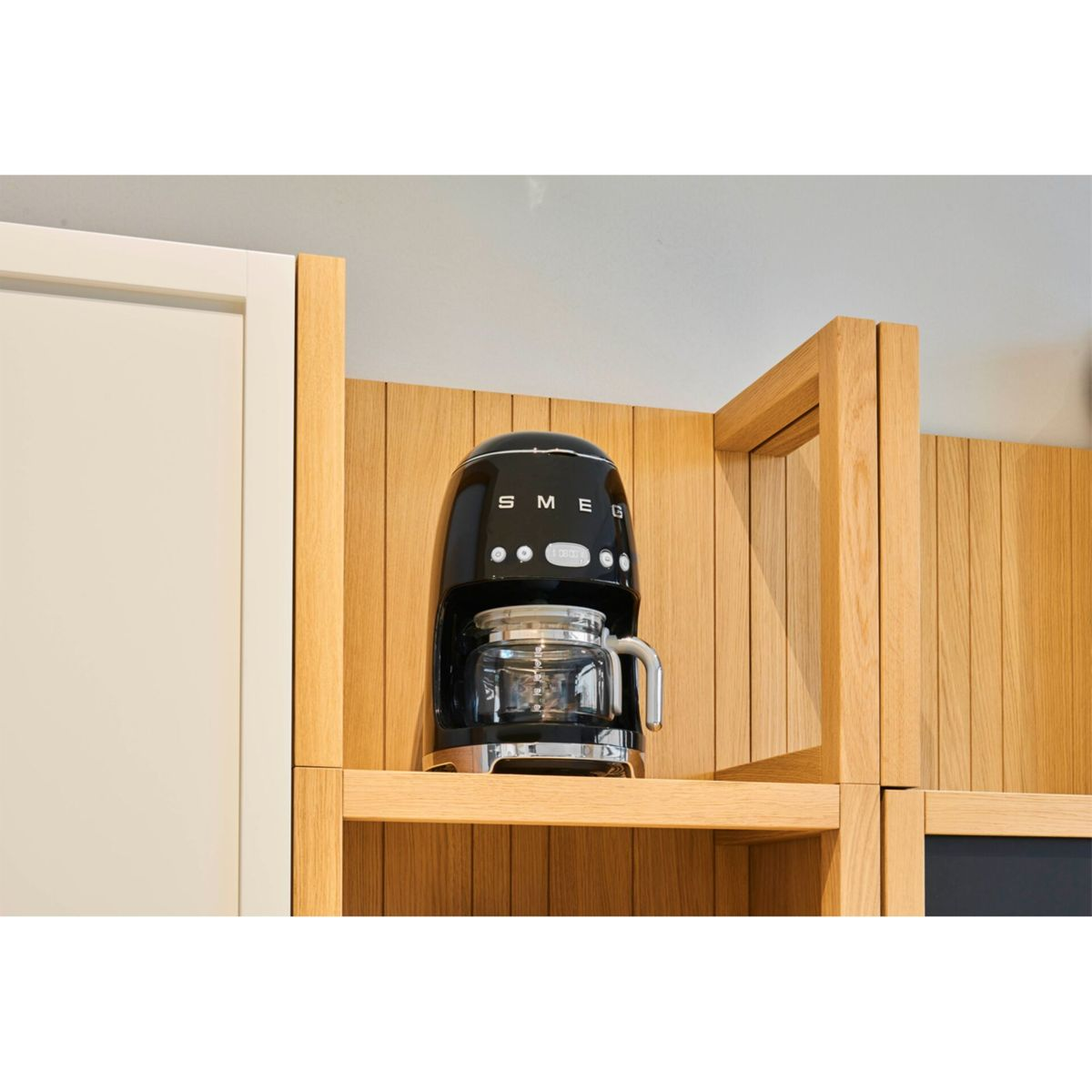 Czarny ekspres do kawy SMEG stoi na drewnianej półce. Ma szklaną karafkę. Tło przedstawia drewniane panele i białą szafkę.