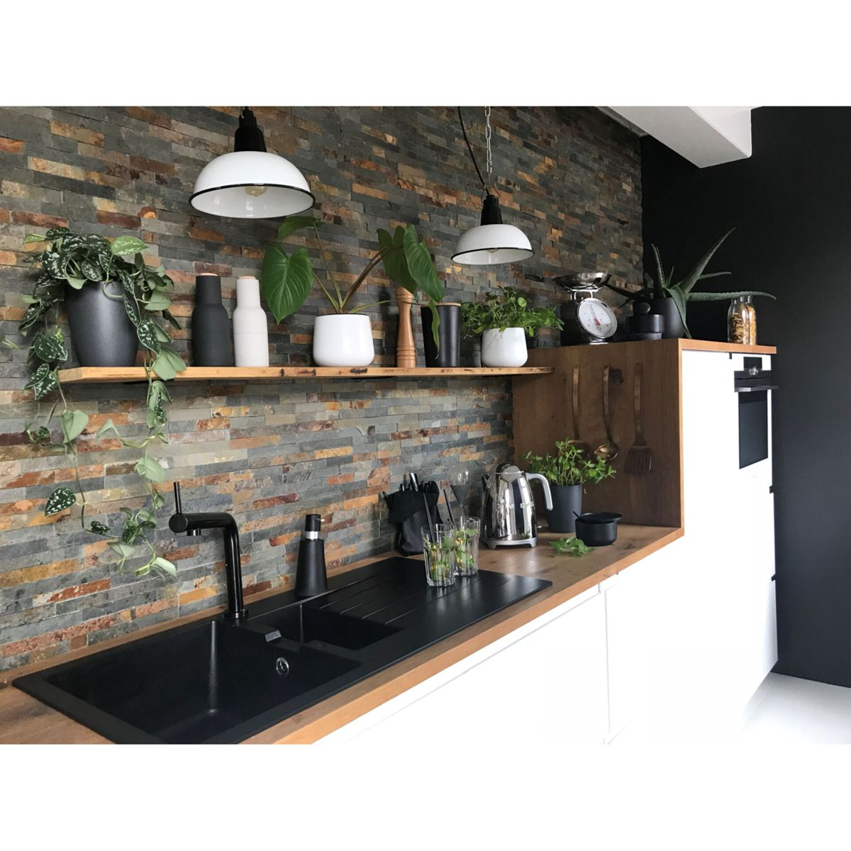 Nowoczesna kuchnia z czarnym zlewem, drewnianym blatem i kamienną ścianą. Rośliny na półkach i blacie.