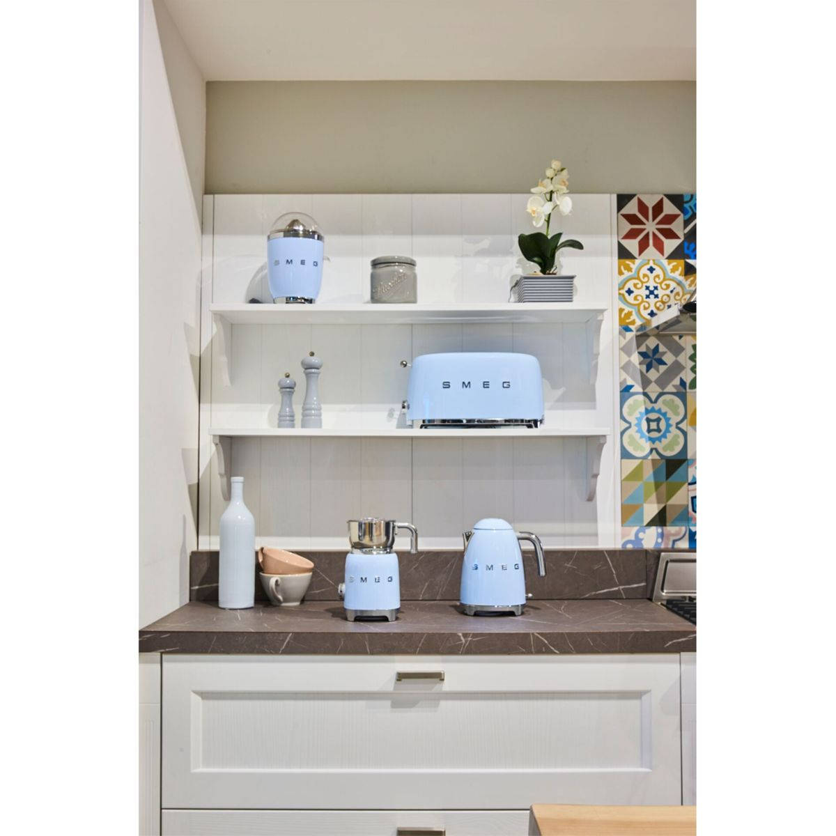 Kuchnia z jasnoniebieskimi urządzeniami SMEG. Toster, blender i czajnik stoją na blacie. Półki zawierają mikser i dekoracje.