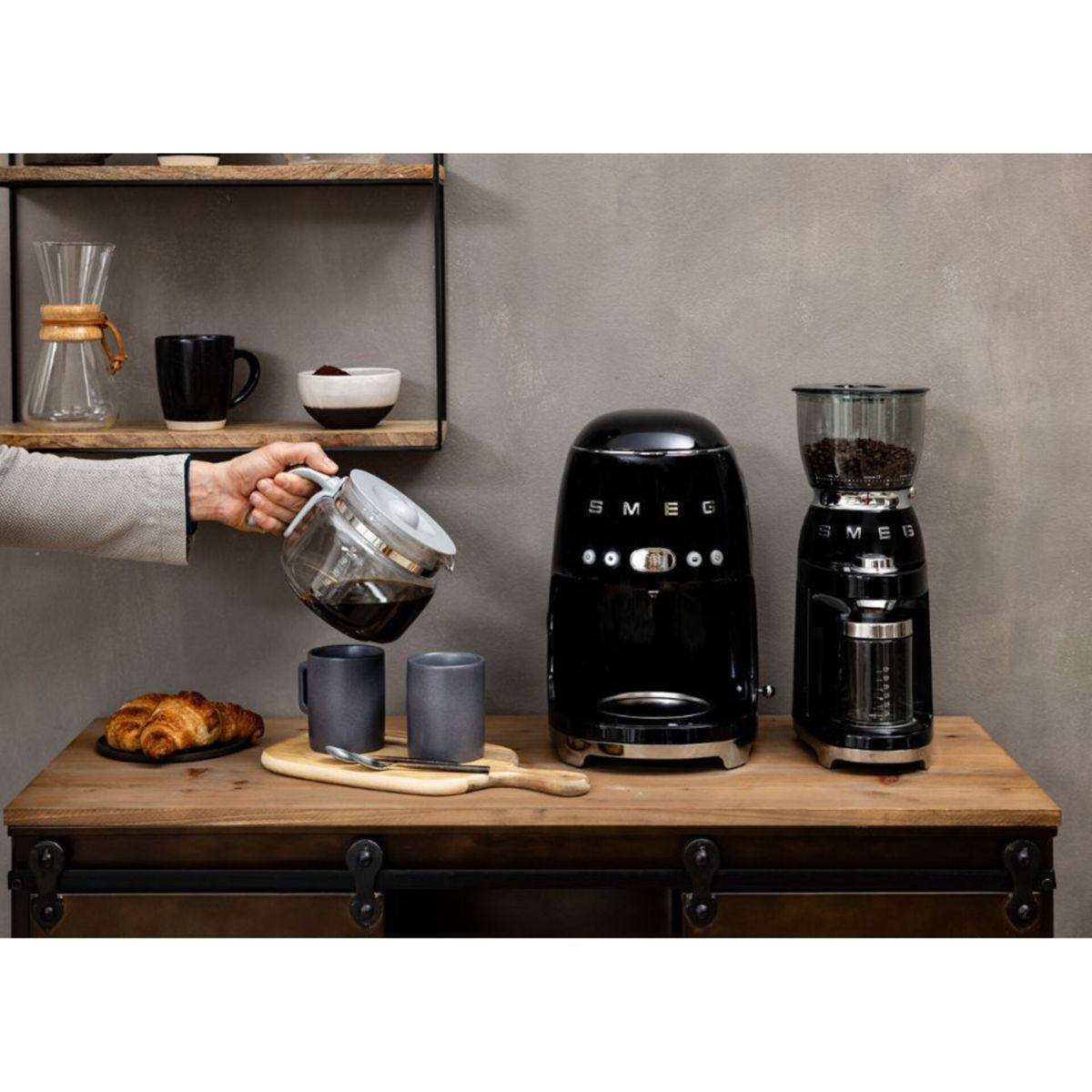 Osoba wlewa kawę ze szklanej karafki do dwóch szarych kubków. Na drewnianym stole stoi czarny ekspres do kawy i młynek.