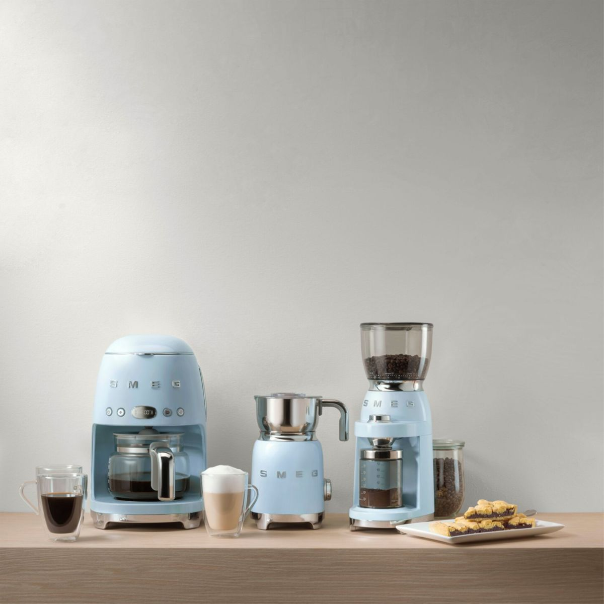 Una macchina da caffè, un macinacaffè e uno schiumatore per latte SMEG azzurri sono su una superficie di legno. Caffè e pasticcini sono accanto.