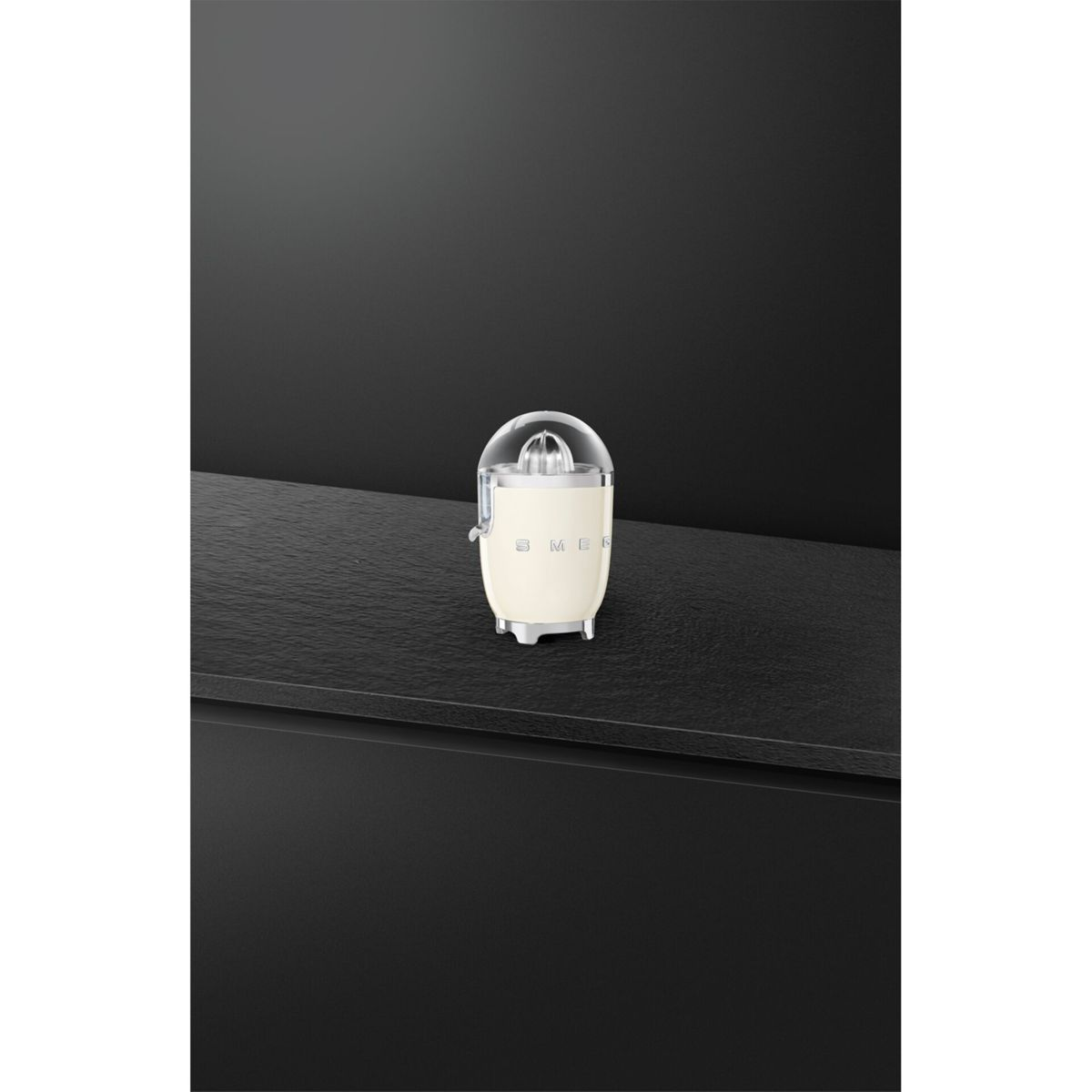 Kremowa wyciskarka do cytrusów Smeg z przezroczystą kopułą na czarnej powierzchni, na ciemnym tle. Wyciskarka jest wyśrodkowana.