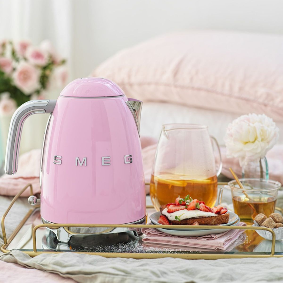 Bouilloire SMEG rose sur un plateau avec du thé, des toasts aux fraises et des fleurs. Fond rose clair avec un oreiller.