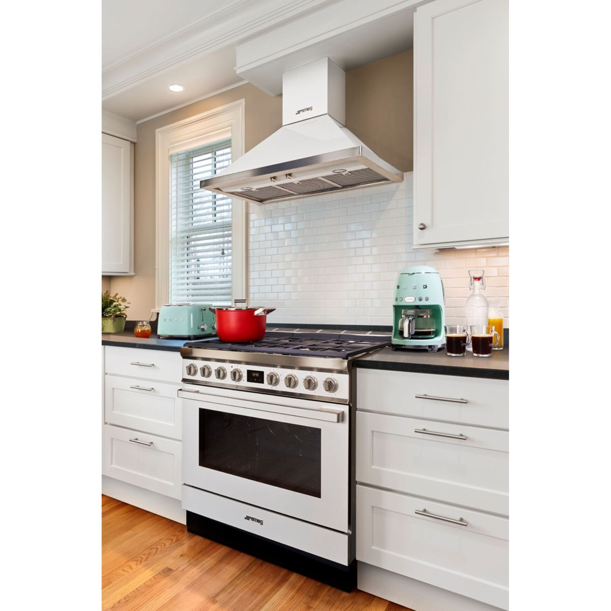 Biała kuchnia z kuchenką, piekarnikiem i okapem. Czerwony garnek na kuchence. Toster i ekspres do kawy widoczne.