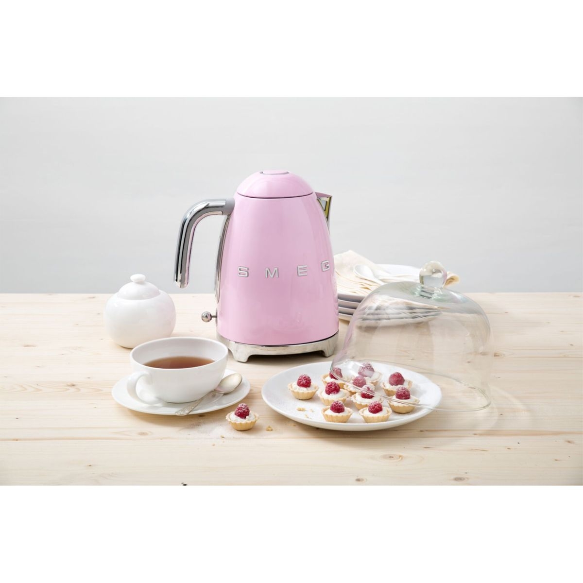 Une bouilloire rose SMEG, une tasse à thé, des pâtisseries sur une assiette, un sucrier et un dôme à gâteau en verre sur une table en bois.