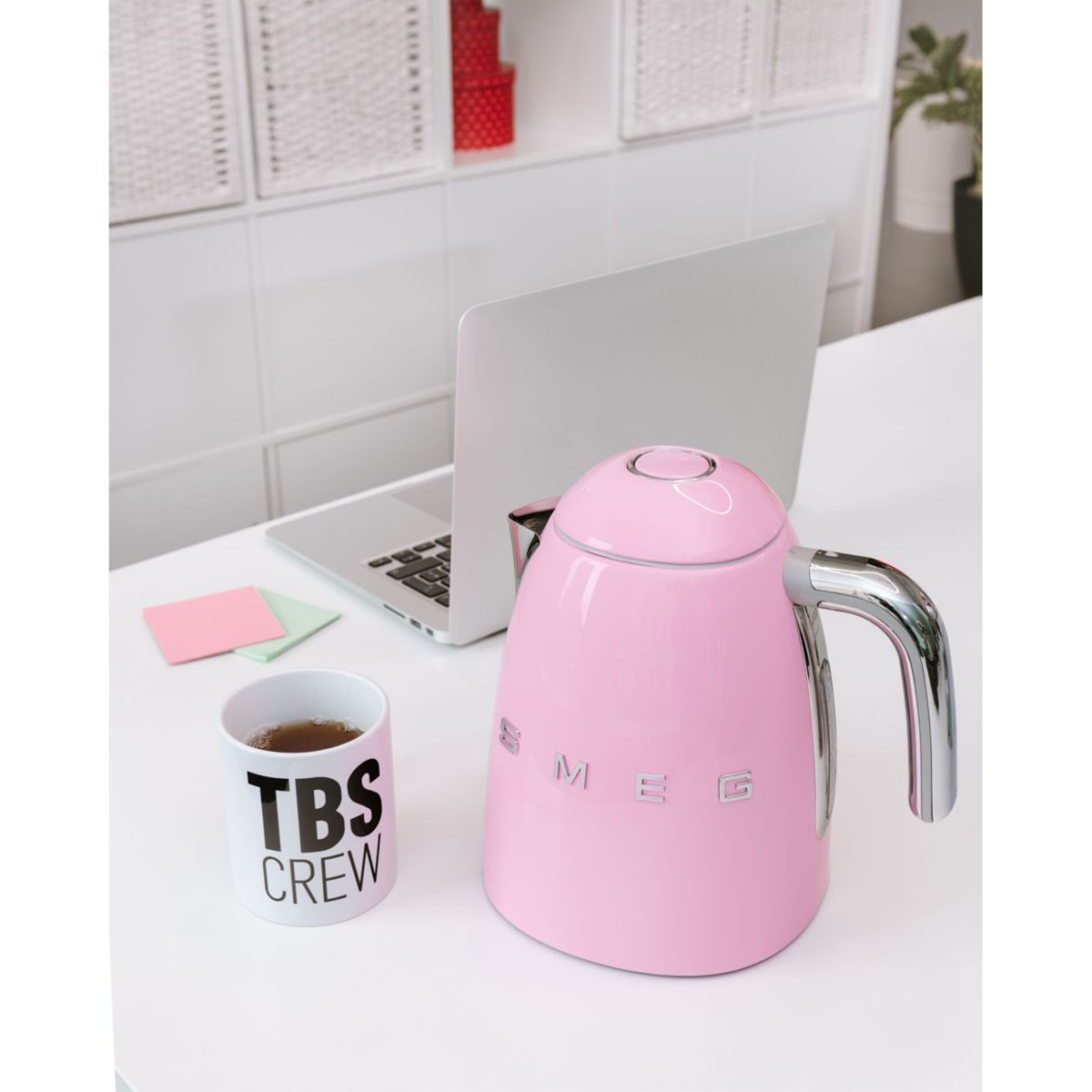 Une bouilloire SMEG rose, un ordinateur portable et une tasse avec 'TBS CREW' sur une table blanche. Le fond a des étagères blanches et une plante.