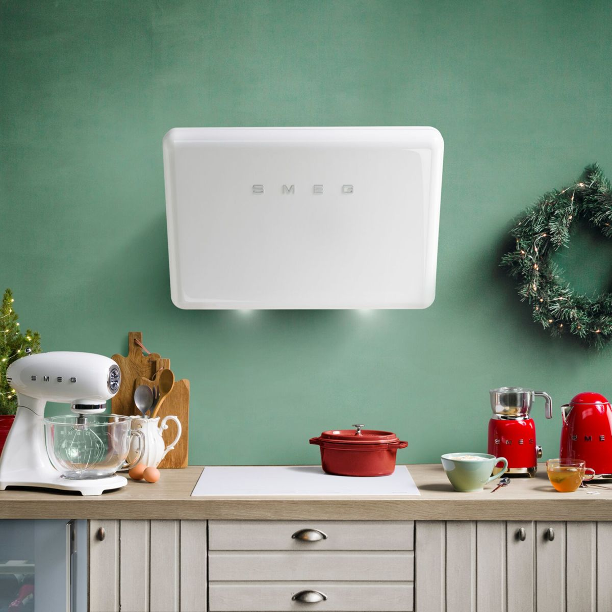 Witte keukenapparatuur op een houten aanrecht tegen een groene muur, inclusief een mixer, een pan en een waterkoker. Een kerstkrans is zichtbaar.