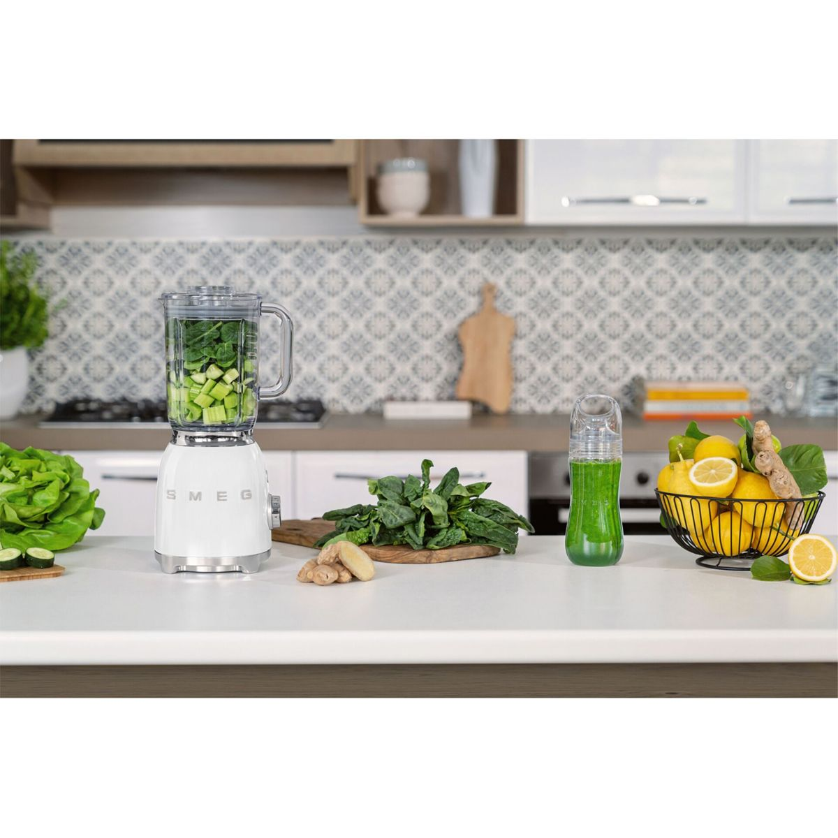 Biały blender SMEG z warzywami, zielonym koktajlem, cytrynami i imbirem na blacie kuchennym. Widoczna jest również sałata i zieleń.