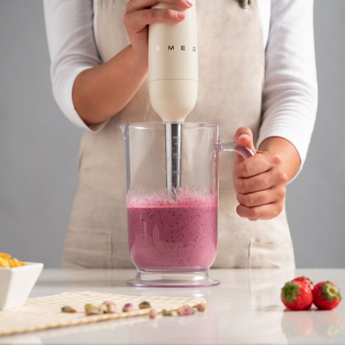 Osoba miksuje różowy koktajl blenderem ręcznym w kolorze kremowym. Składniki i truskawki na stole.