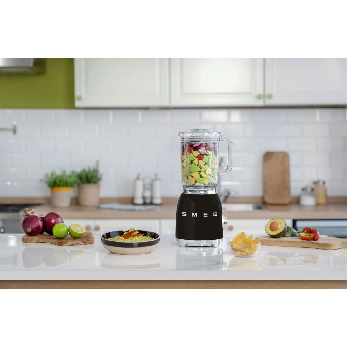 Czarny blender Smeg wypełniony warzywami stoi na białym blacie. Obok miska z jedzeniem, chipsami i składnikami.