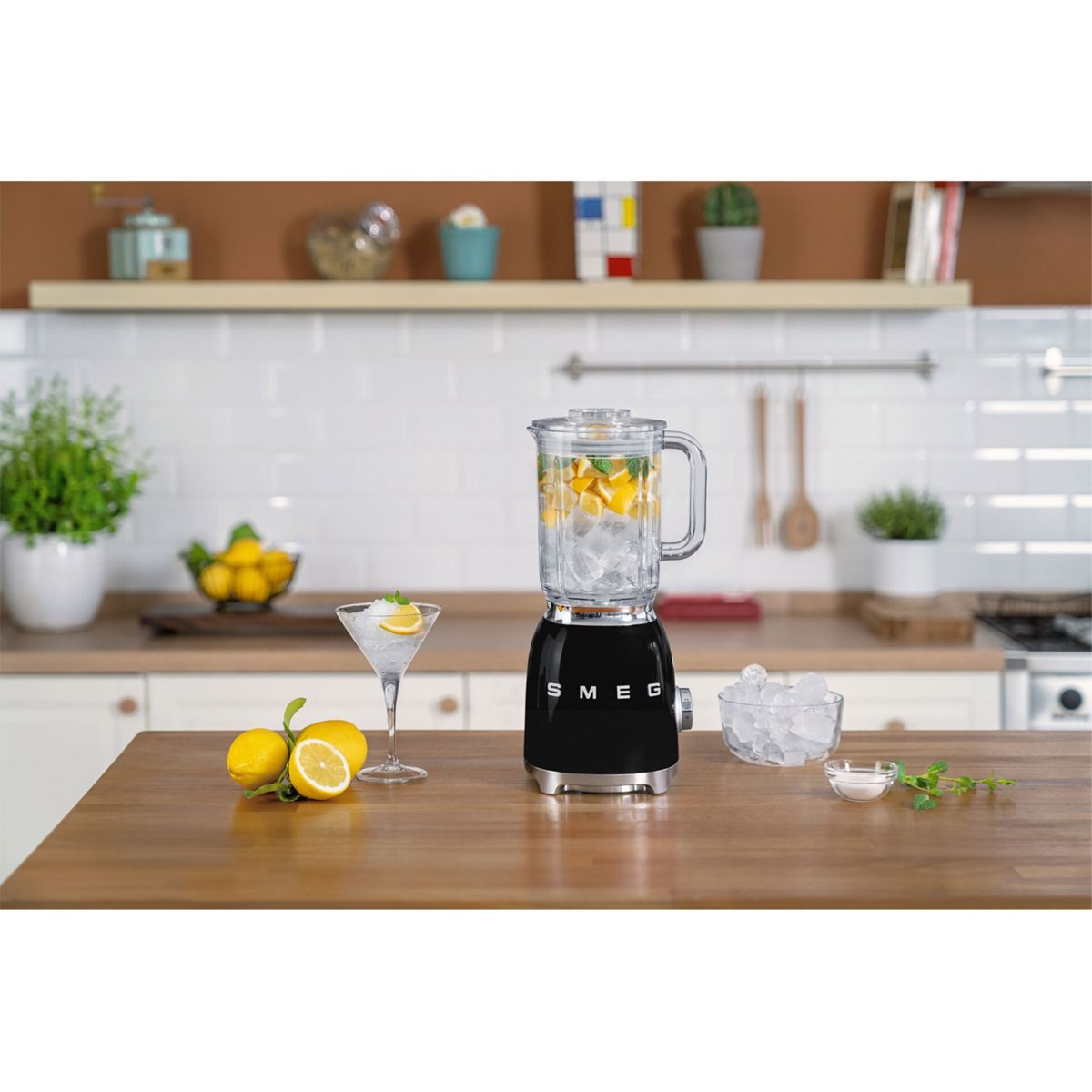 Czarny blender SMEG z lodem, cytrynami i kieliszkiem do koktajli na drewnianym blacie. Tło kuchni z roślinami, cytrynami i przyborami.
