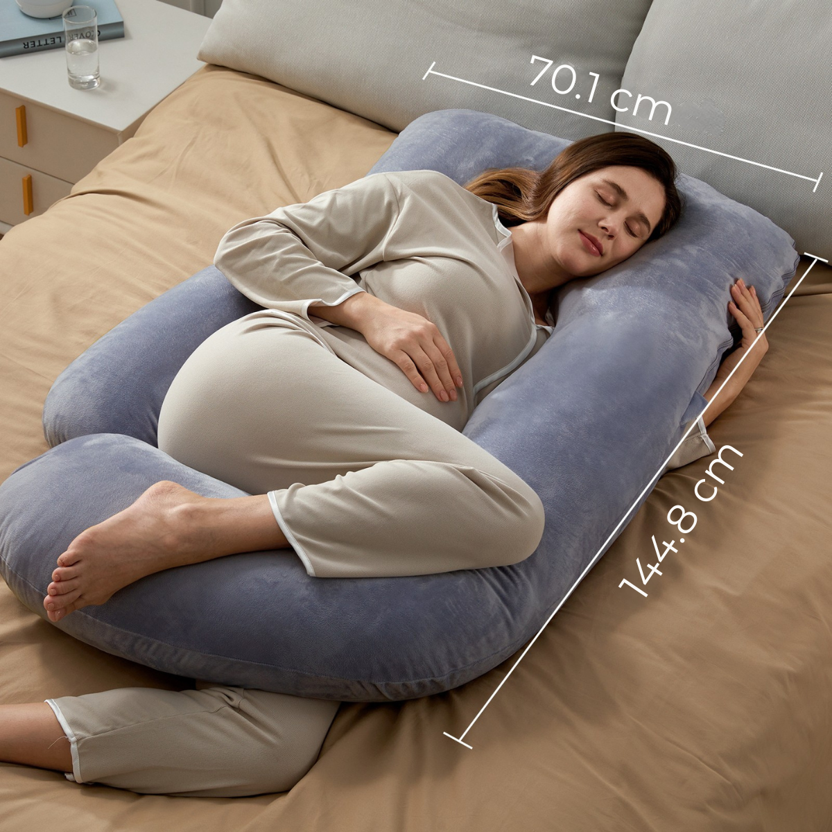 Zwangere vrouw ligt op een U-vormig grijs kussen op een bed. Ze draagt beige pyjama. Het beddengoed is bruin. Afmetingen zijn aangegeven.