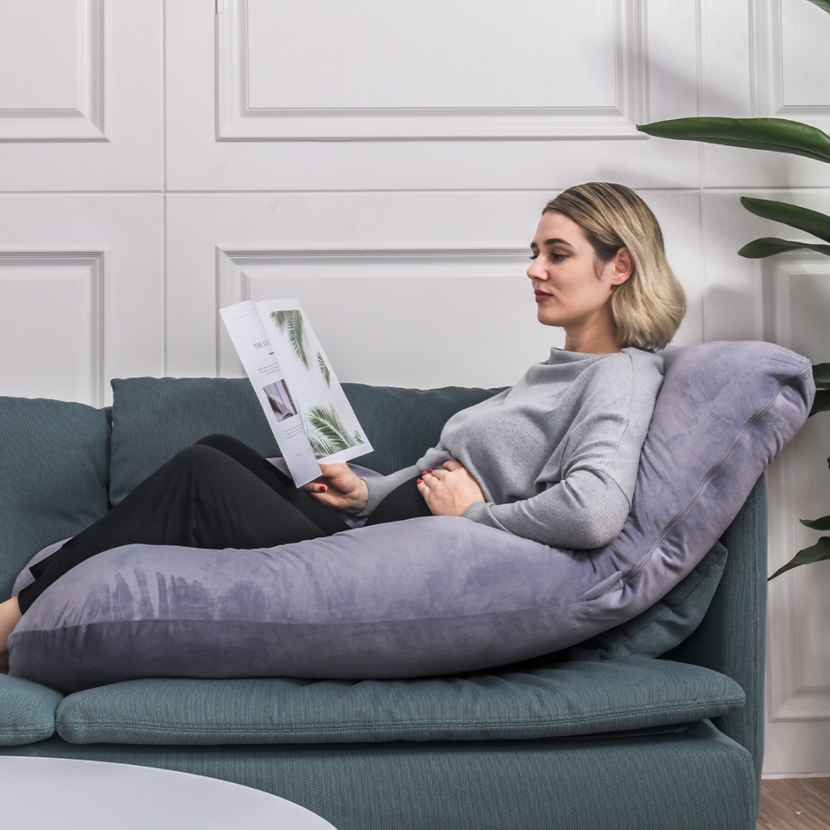 Een vrouw leest een tijdschrift op een blauwgroene bank, ondersteund door een groot grijs kussen. Ze draagt een grijze trui en een zwarte broek.