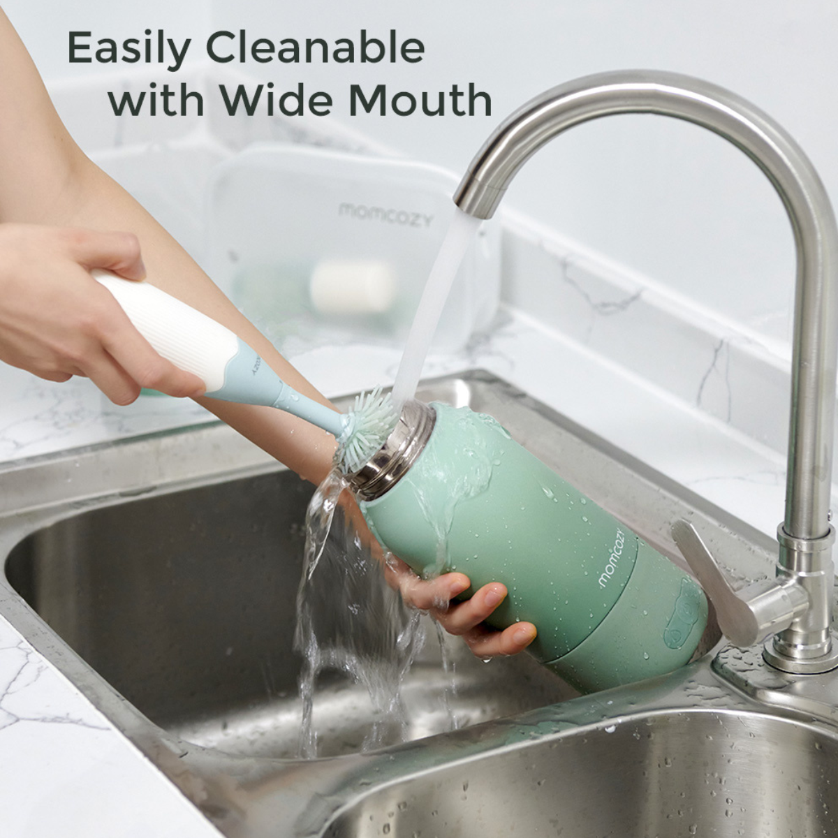 A person is cleaning a green water bottle under running water in a stainless steel sink, using a brush.