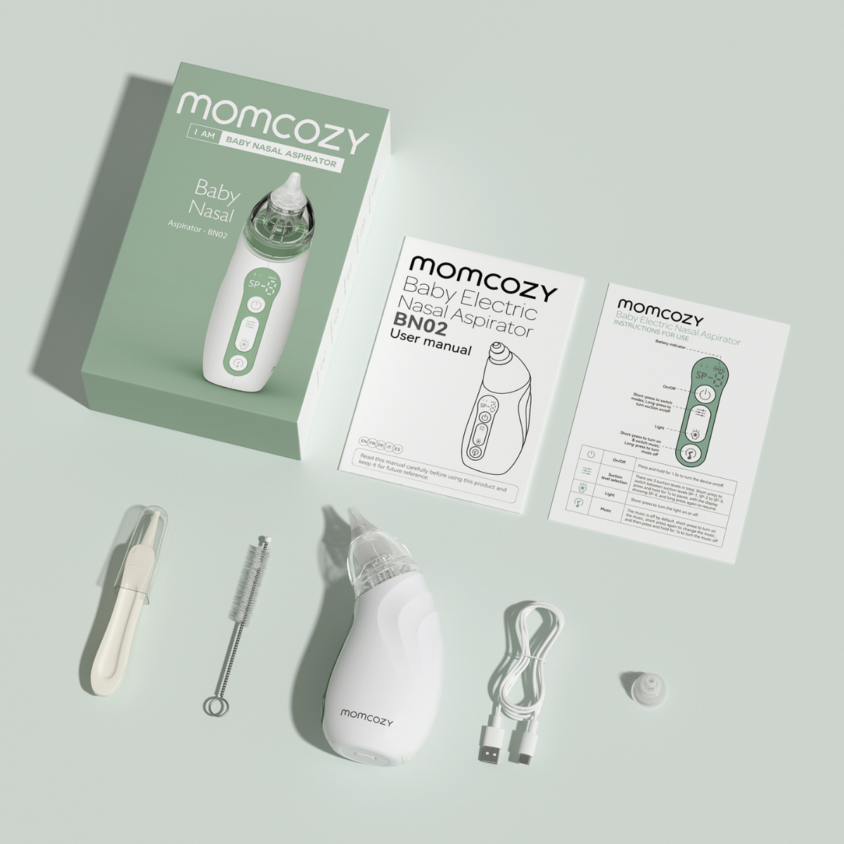 A white and green Momcozy baby nasal aspirator kit is displayed with manuals, cleaning tools, and a charging cable on a light green surface.