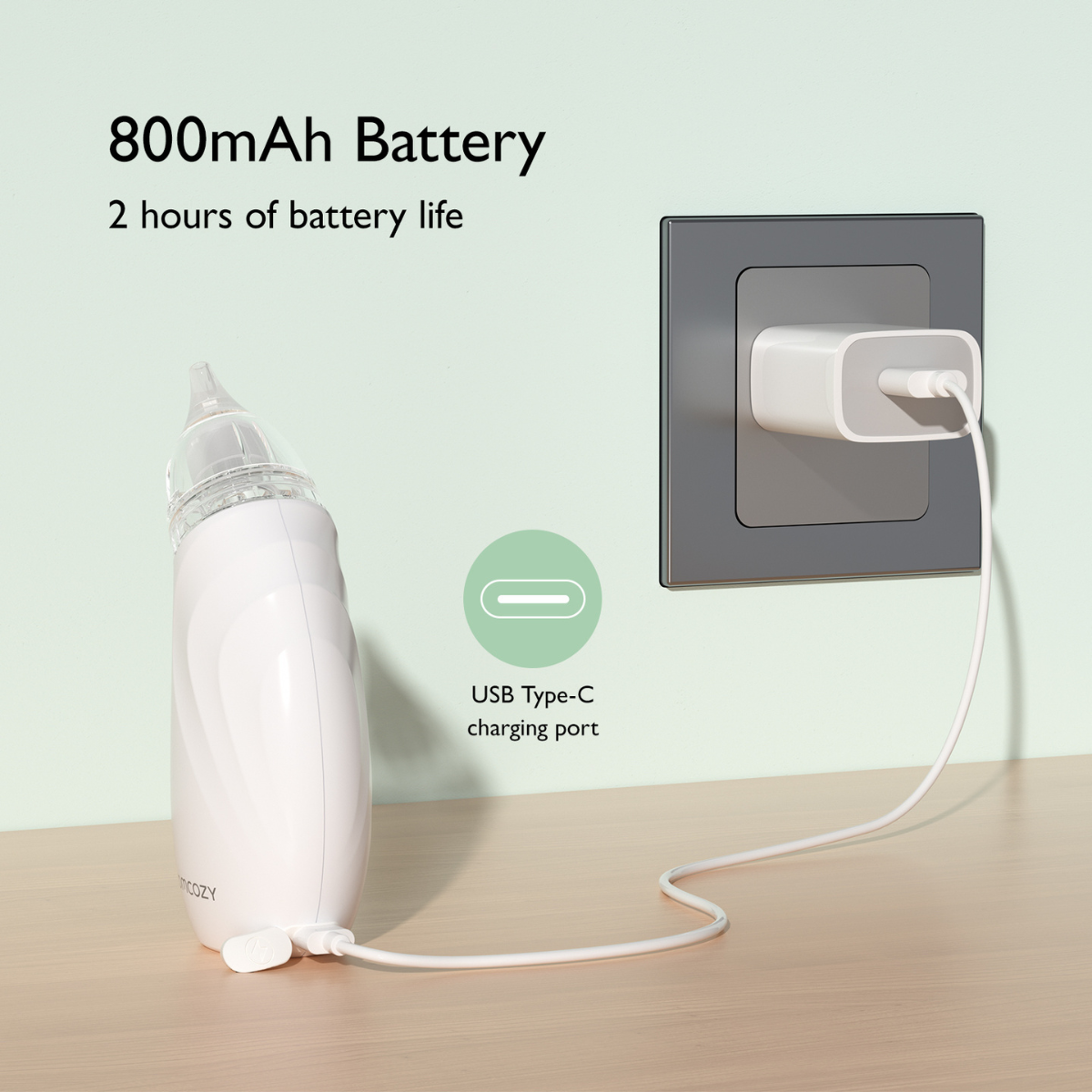 A white baby nasal aspirator is charging. A white charger is plugged into a wall. The aspirator is on a wooden surface.