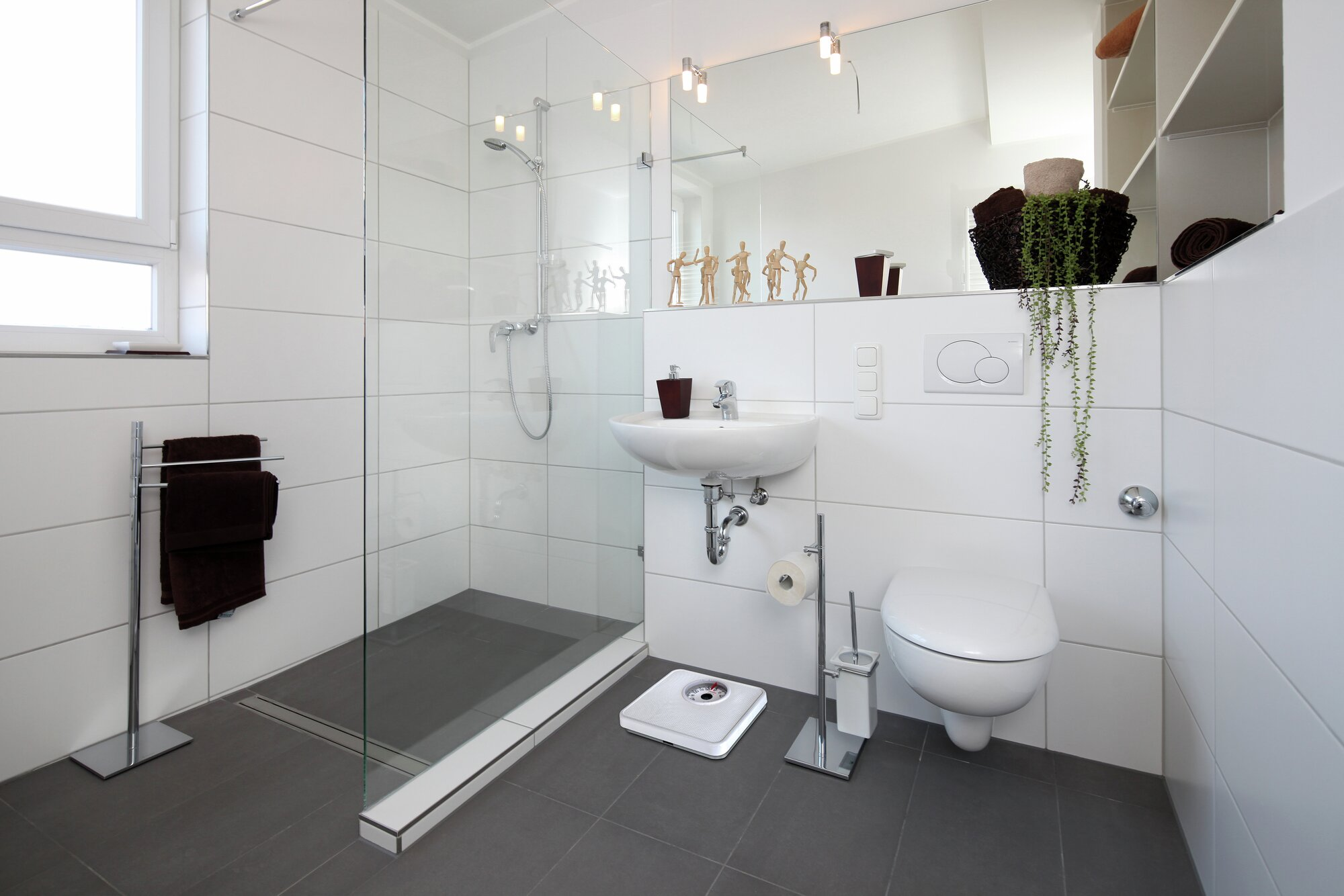 Nowoczesna łazienka z białymi kafelkami, szklanym prysznicem, toaletą, umywalką i wagą na szarej podłodze z płytek.