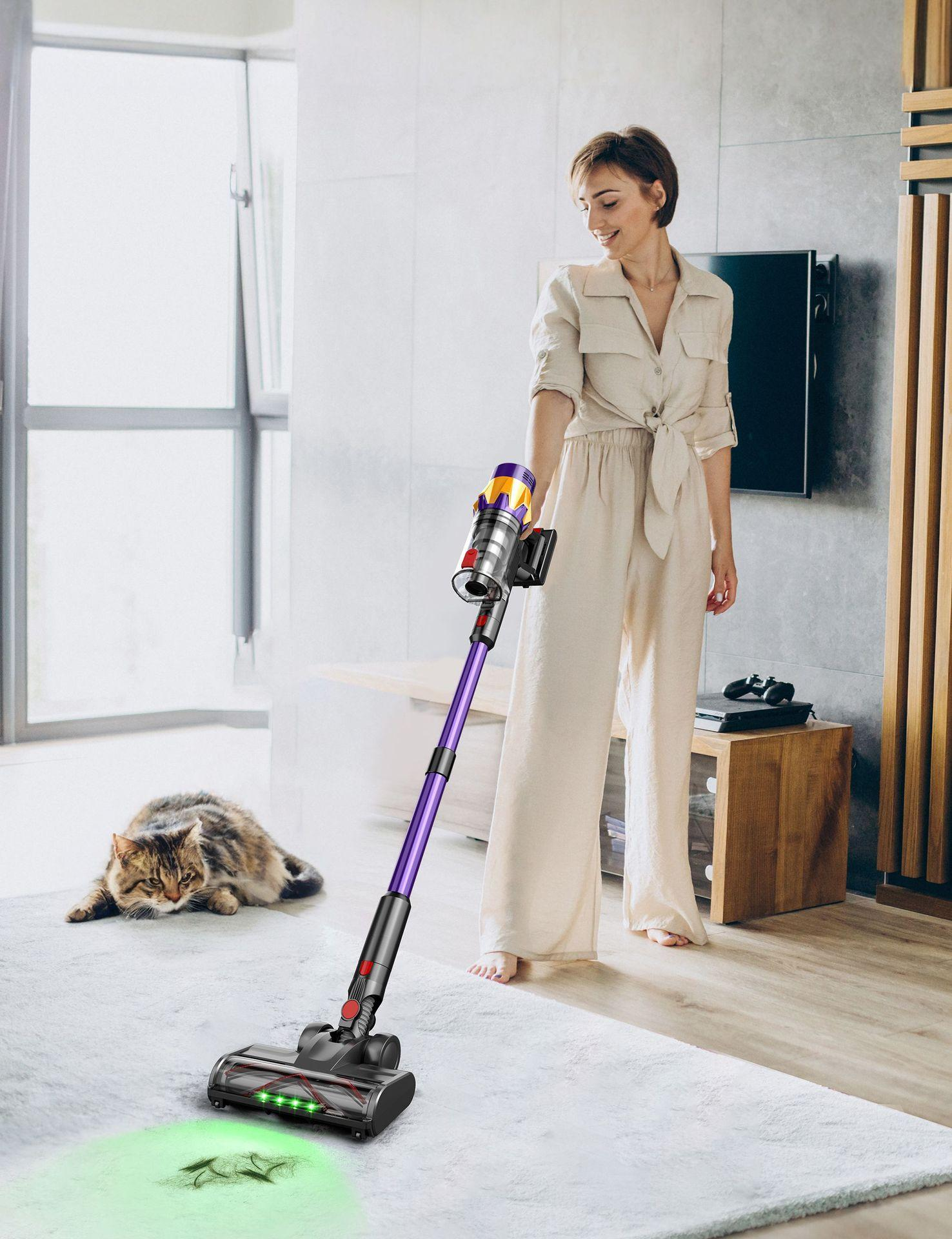 Eine Frau saugt einen Teppich mit einer Katze. Der Staubsauger ist lila und silber. Die Frau trägt ein beiges Outfit. Ein Fernseher und eine Holzverkleidung sind im Hintergrund.