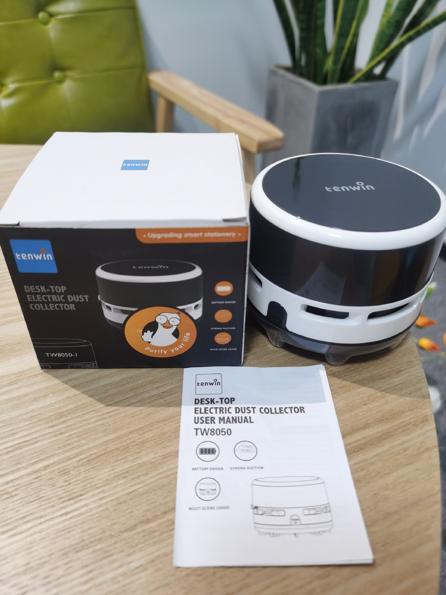 Ein elektrischer Tischstaubsauger mit Box und Bedienungsanleitung liegt auf einem Holztisch. Eine Pflanze in einem grauen Topf ist im Hintergrund.