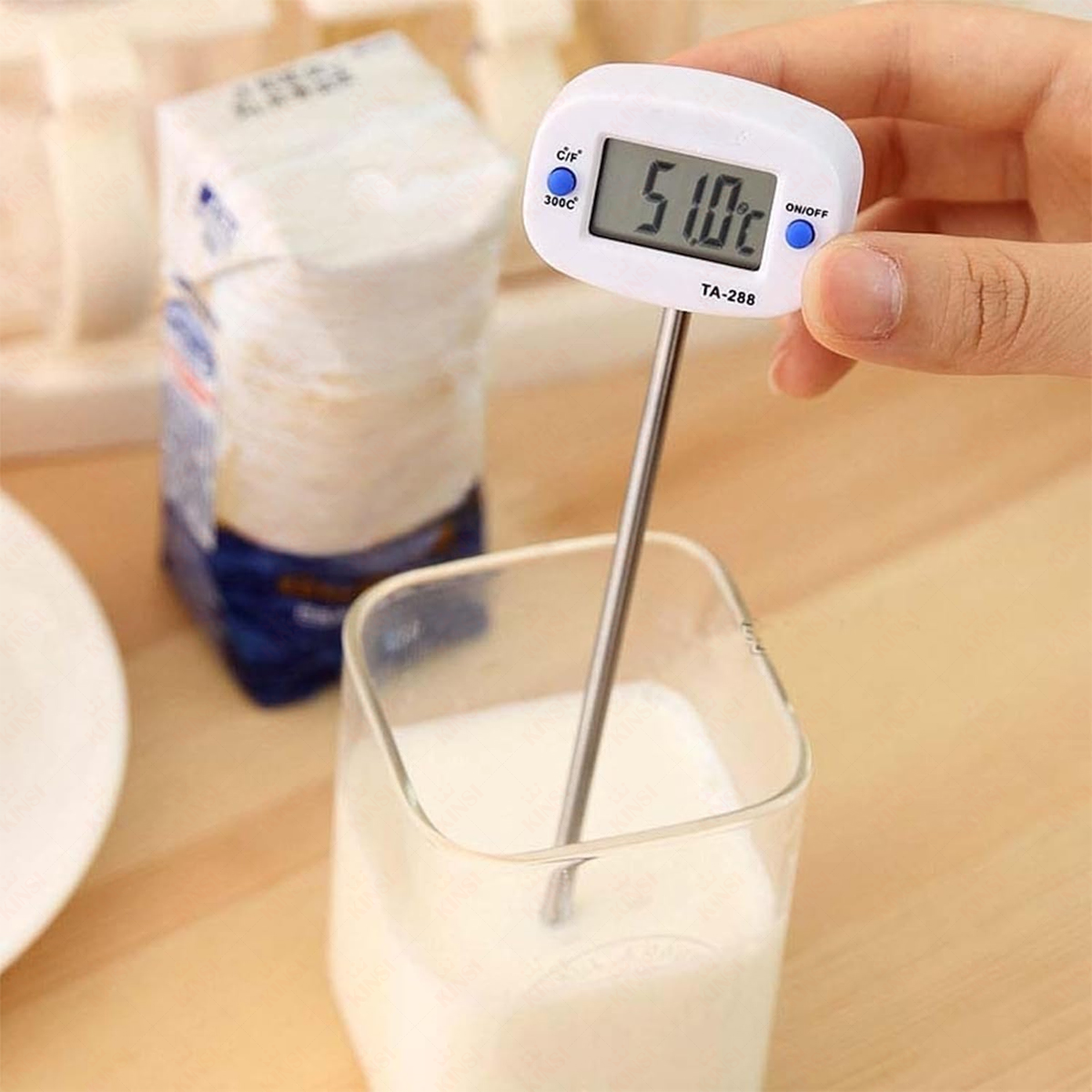 Eine Hand hält ein digitales Thermometer in Milch. Hinter einem Glas Milch steht ein Milchkarton. Das Thermometer zeigt 51,0 °C an.
