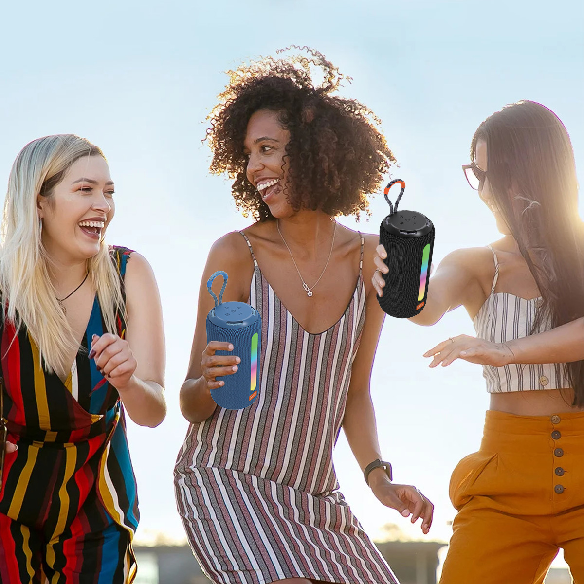 Trzy kobiety śmieją się i trzymają głośniki. Są na zewnątrz w słoneczny dzień. Jeden głośnik jest niebieski, a drugi czarny.