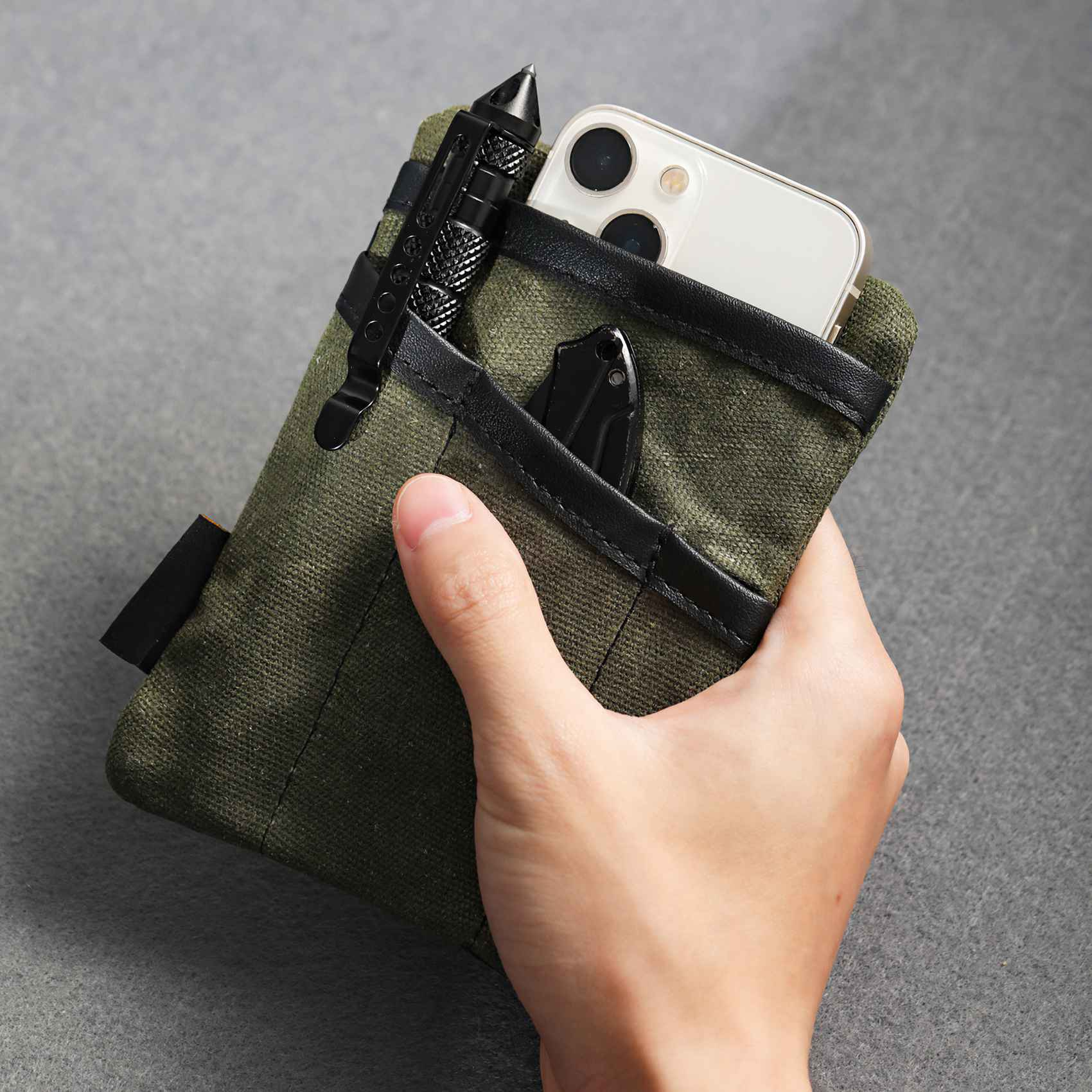 Hand hält einen olivgrünen Beutel mit einem Telefon, einem Messer und einem schwarzen stiftartigen Werkzeug. Der Hintergrund ist eine graue Oberfläche.