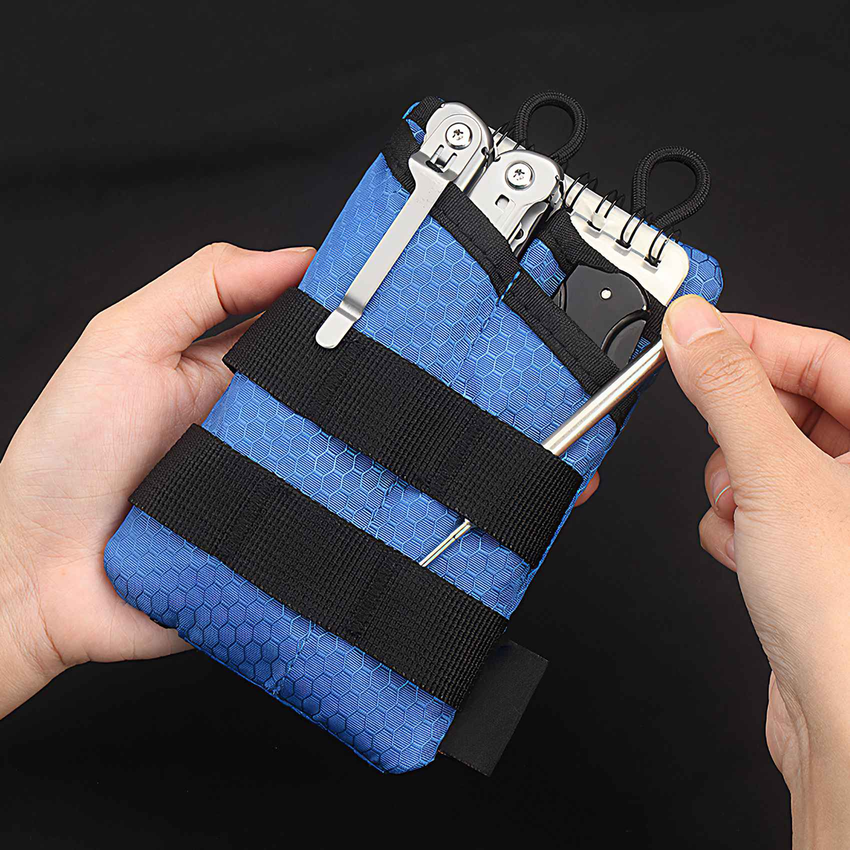 Hände halten eine blaue Tasche mit Werkzeugen, darunter ein Messer, ein Multitool und ein Notizblock, vor schwarzem Hintergrund.