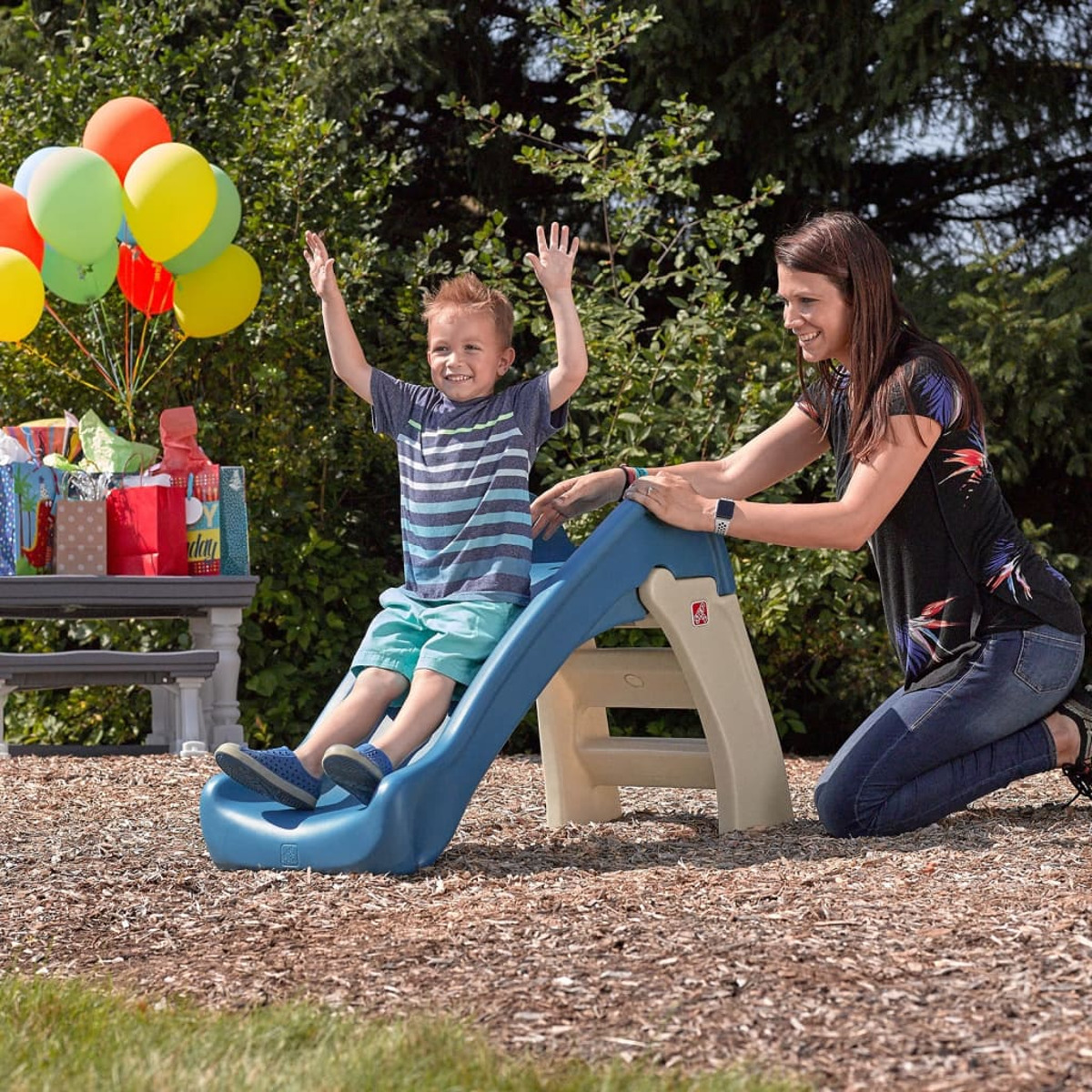 Ein Junge rutscht eine blaue Rutsche hinunter, während eine Frau zusieht. Luftballons und Geschenke sind im Hintergrund zu sehen, was auf eine Party hindeutet.